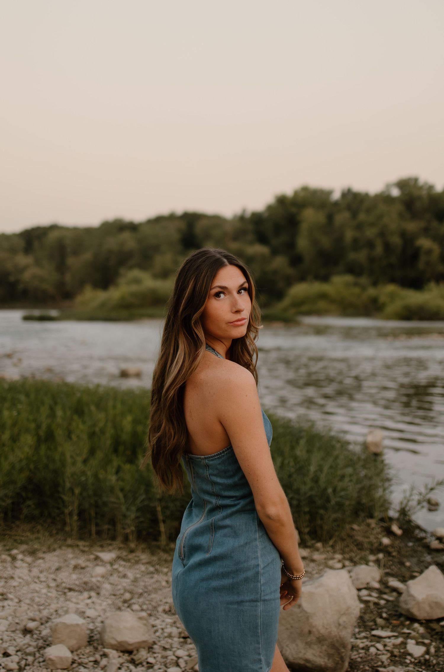 Young woman in denim dress poses by a scenic river at golden hour with lush green trees in background.