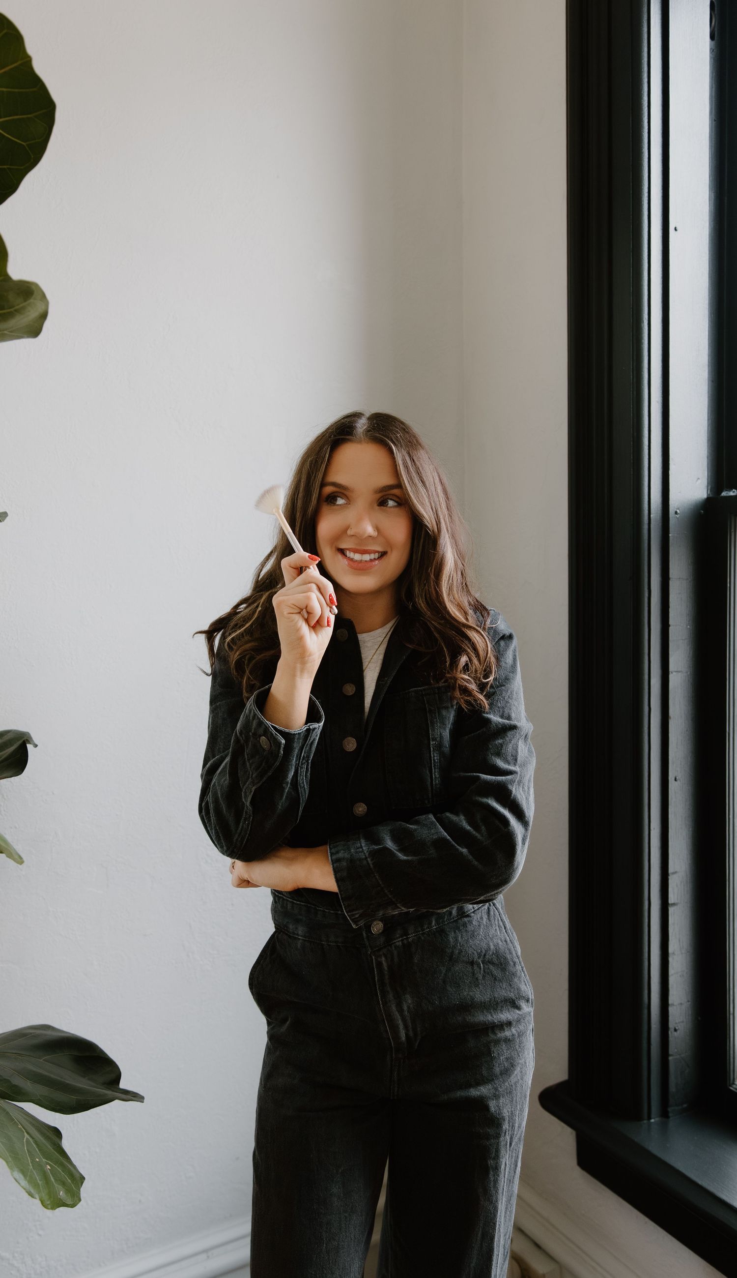 Smiling woman in dark denim suit holding pen near window with green plants.