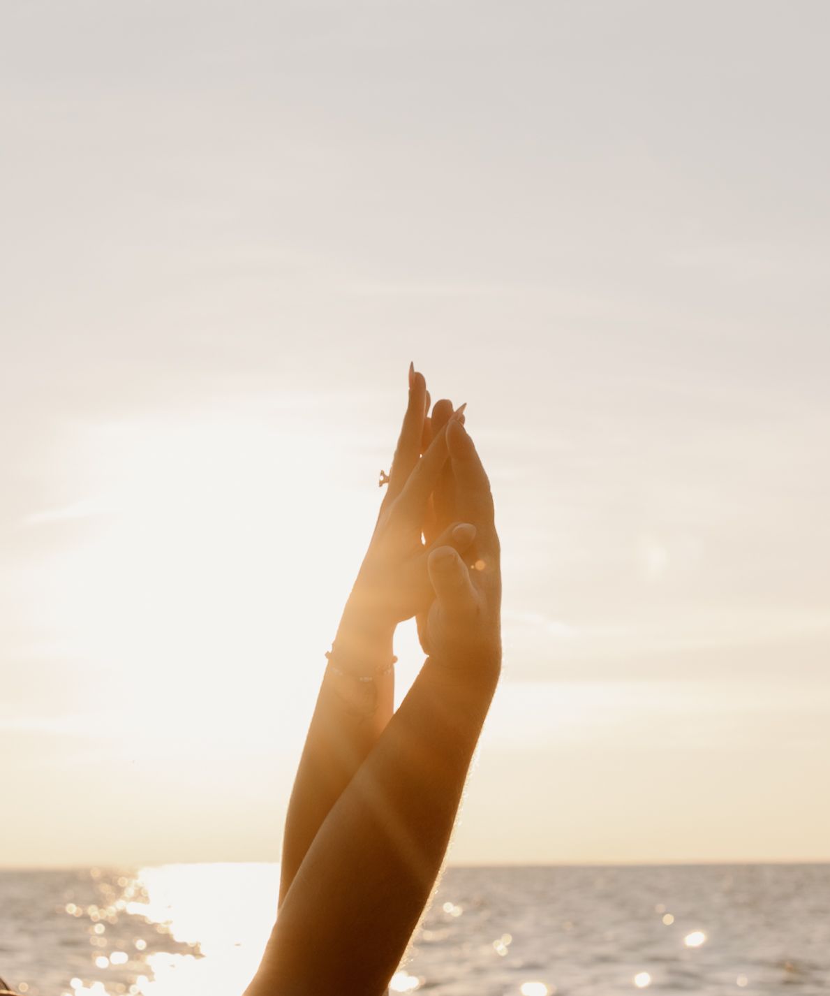 Two hands reaching toward the golden sunset sky over a calm ocean, adorned with delicate rings.