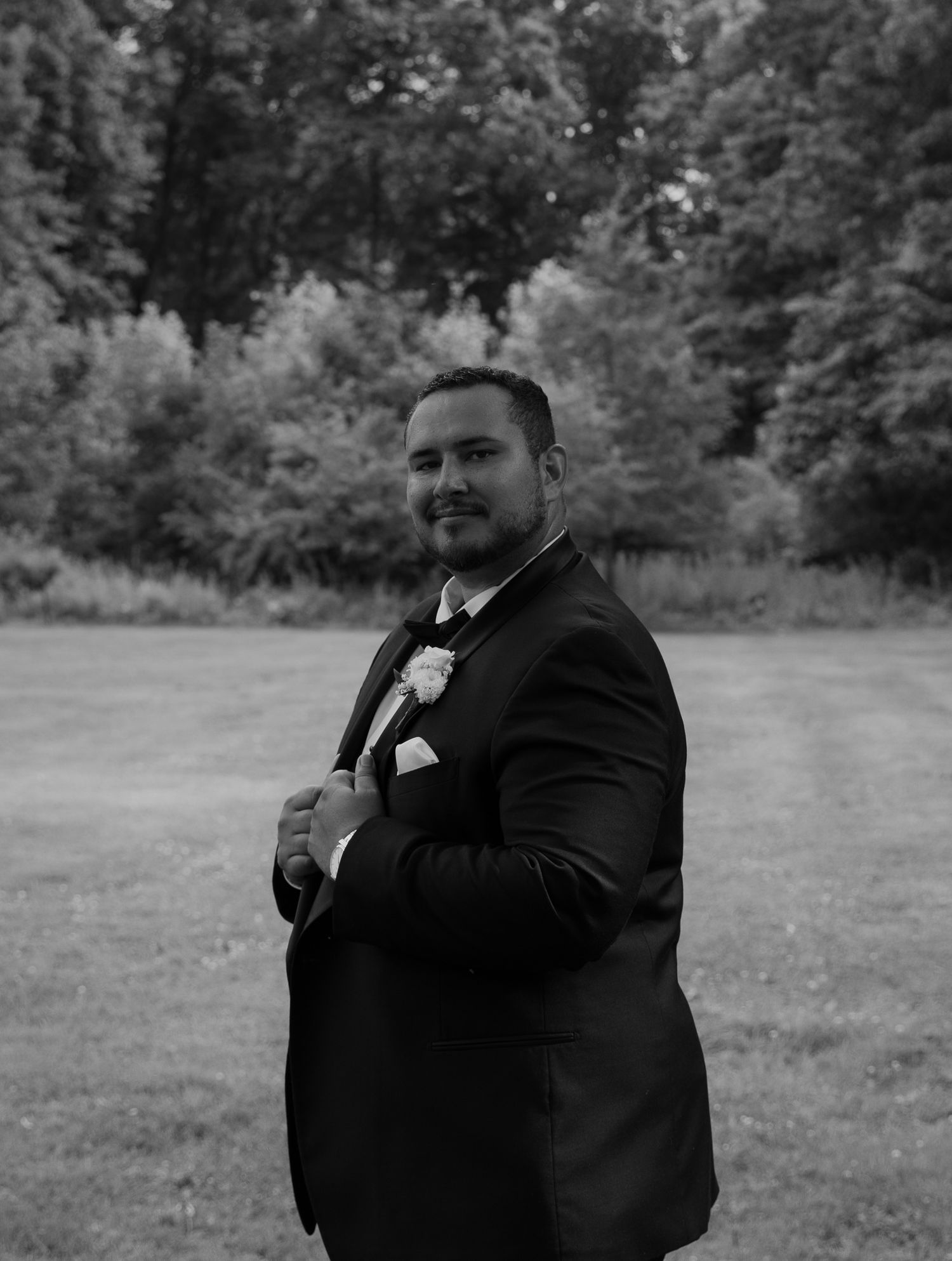 Dapper groom in black tuxedo with boutonniere poses outdoors in black and white wedding photo.