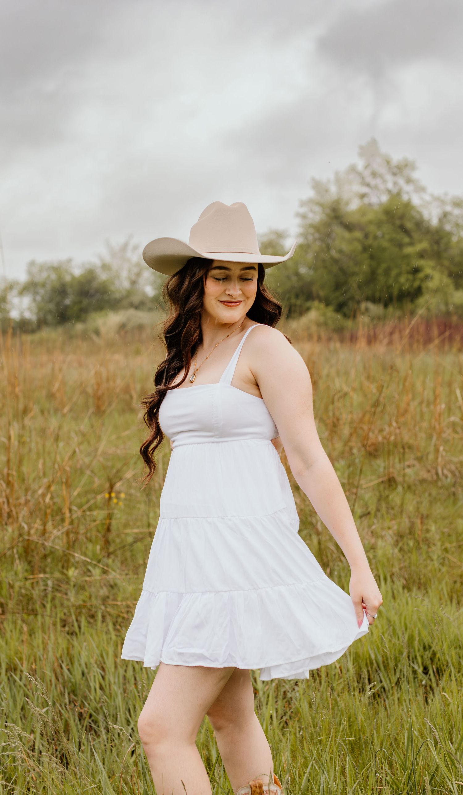 Woman in white sundress and beige cowboy hat poses in rural field with tall grass on cloudy day.