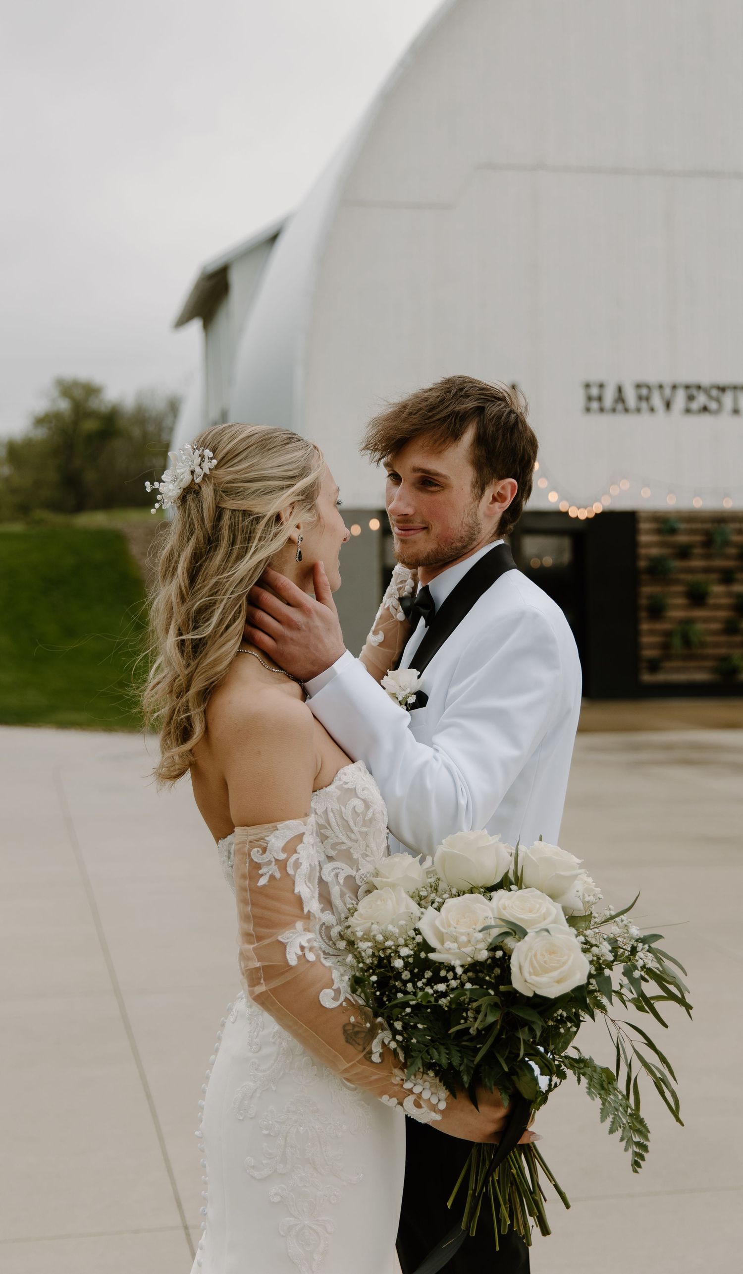 A bride and groom share a romantic moment outside a white barn wedding venue, bride holding white roses bouquet.