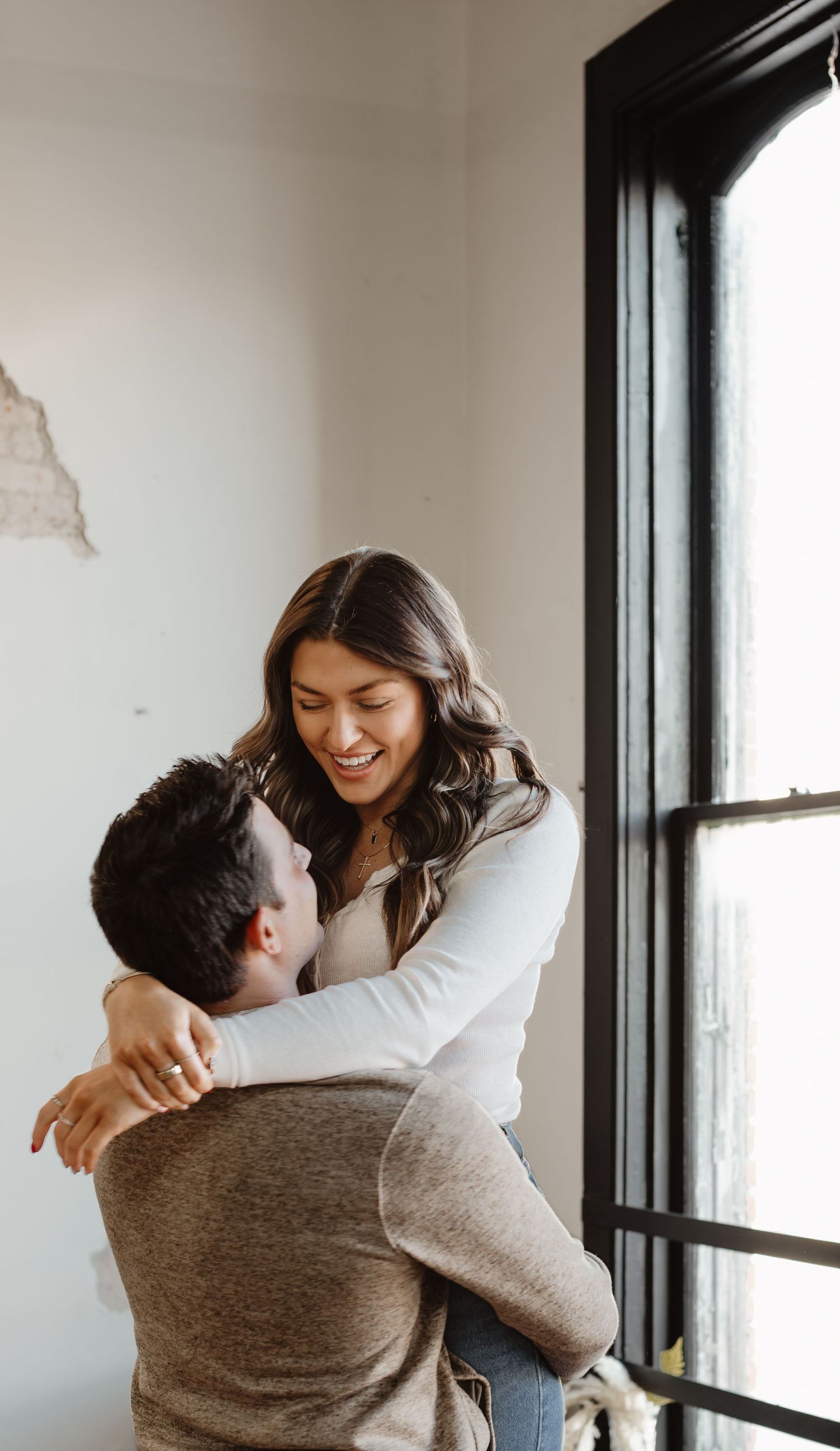 Couple sharing a joyful embrace by a large window in an industrial-style space.