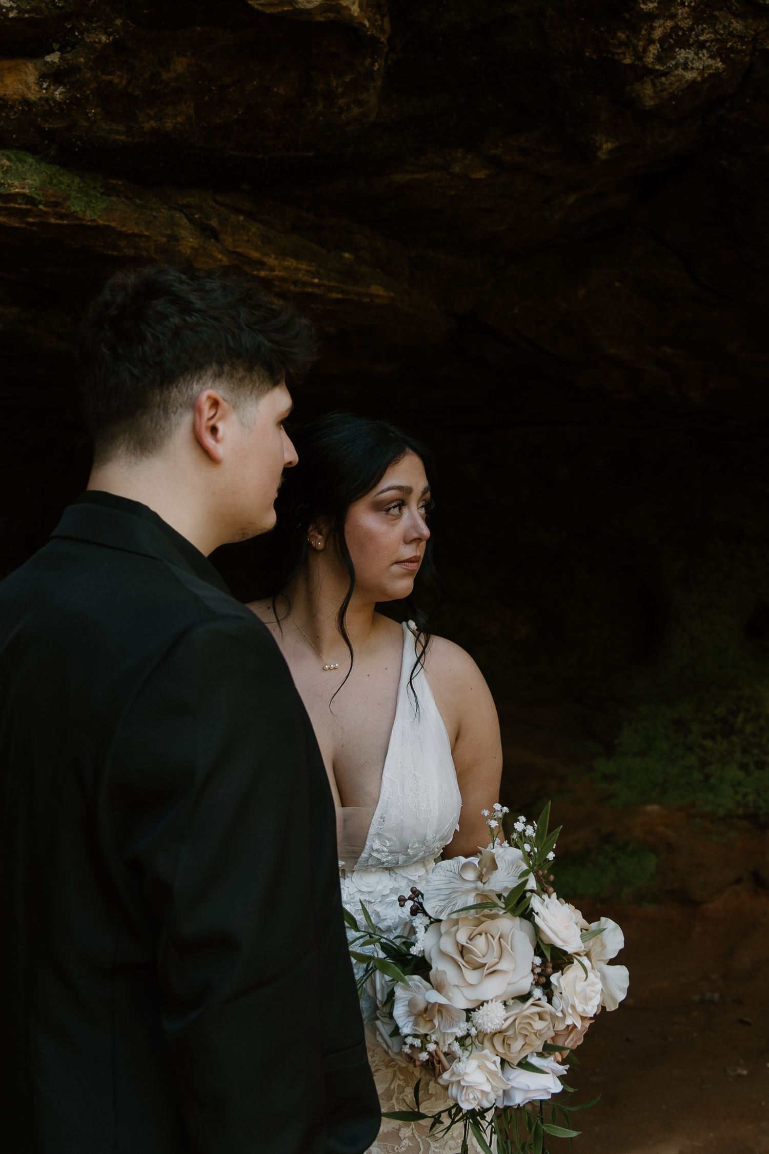 A bride holding white floral bouquet stands beside groom in black suit near dark rocky cave backdrop.