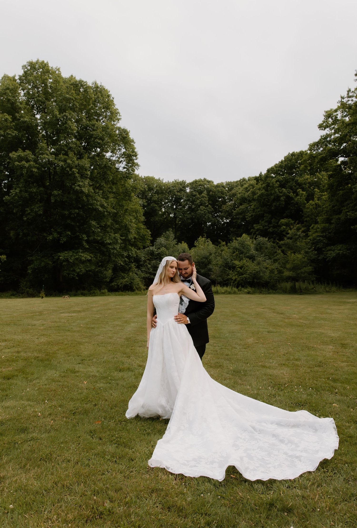 Bride and groom embrace in a lush green field, her lace wedding gown train flowing elegantly on the grass.