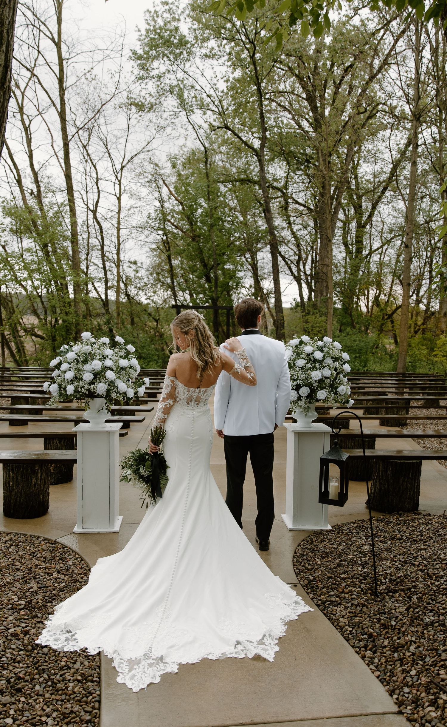 Bride and groom walk down outdoor wedding aisle lined with white floral arrangements and wooden benches.