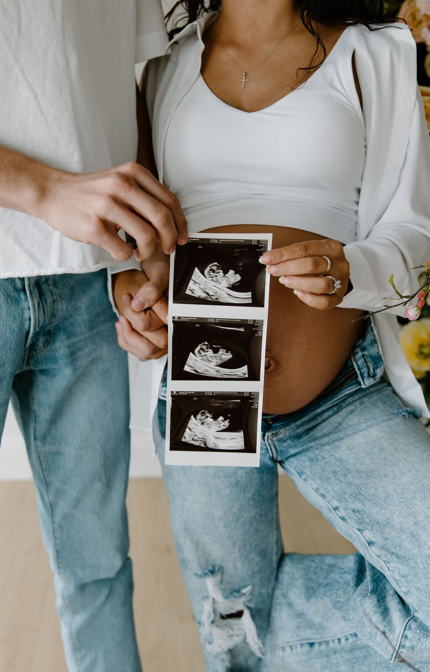 Pregnant couple holding ultrasound sonogram photos showing baby bump in white crop top and ripped jeans.
