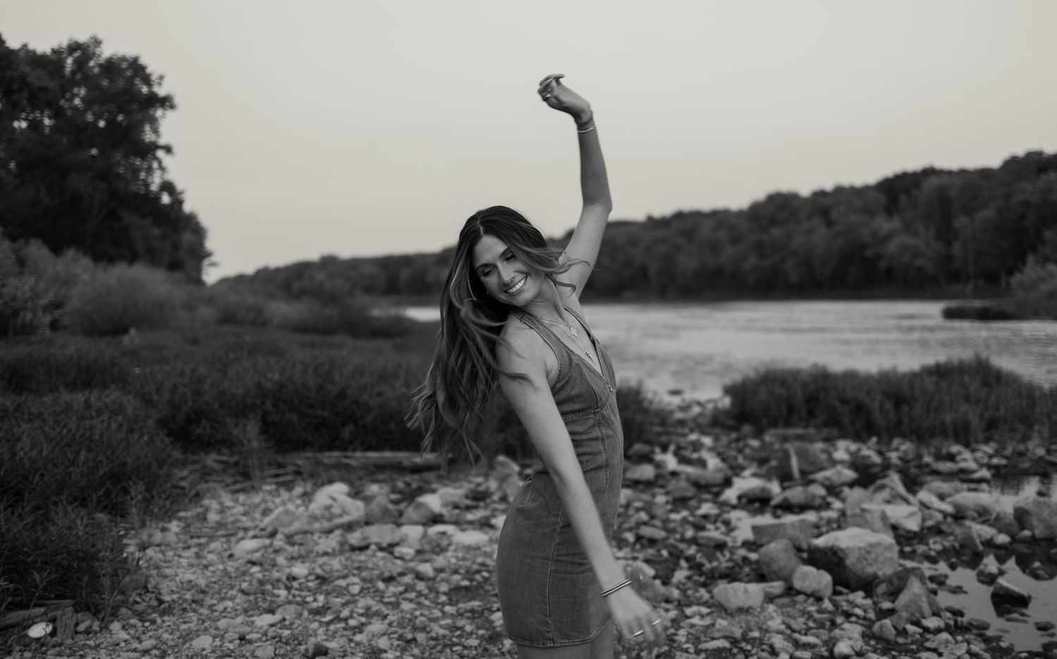 Black and white photo of a woman posing outdoors near a rocky riverbank, raising her arm gracefully.