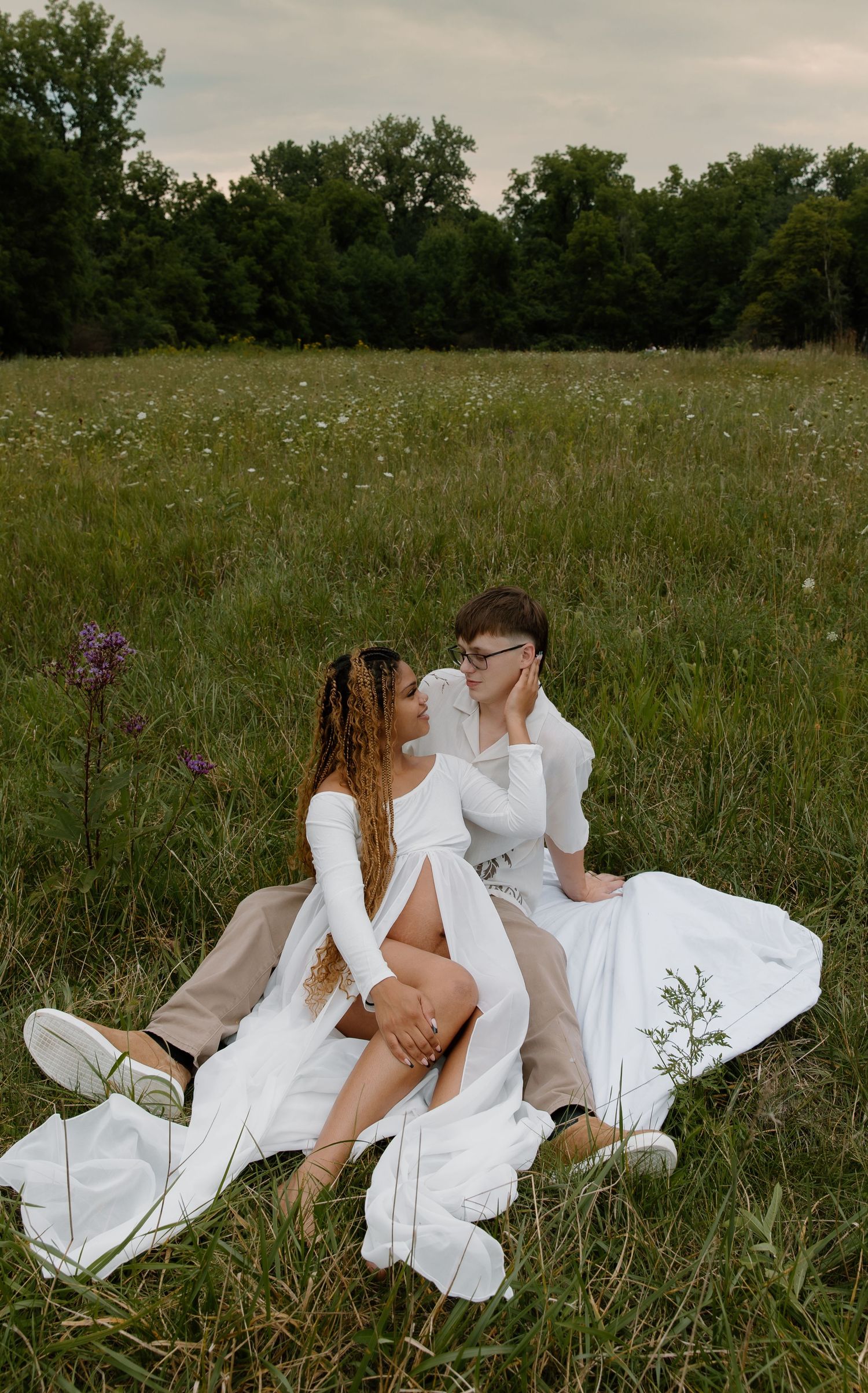 Couple in white outfits sitting romantically in a wildflower meadow with lush green trees in the background.