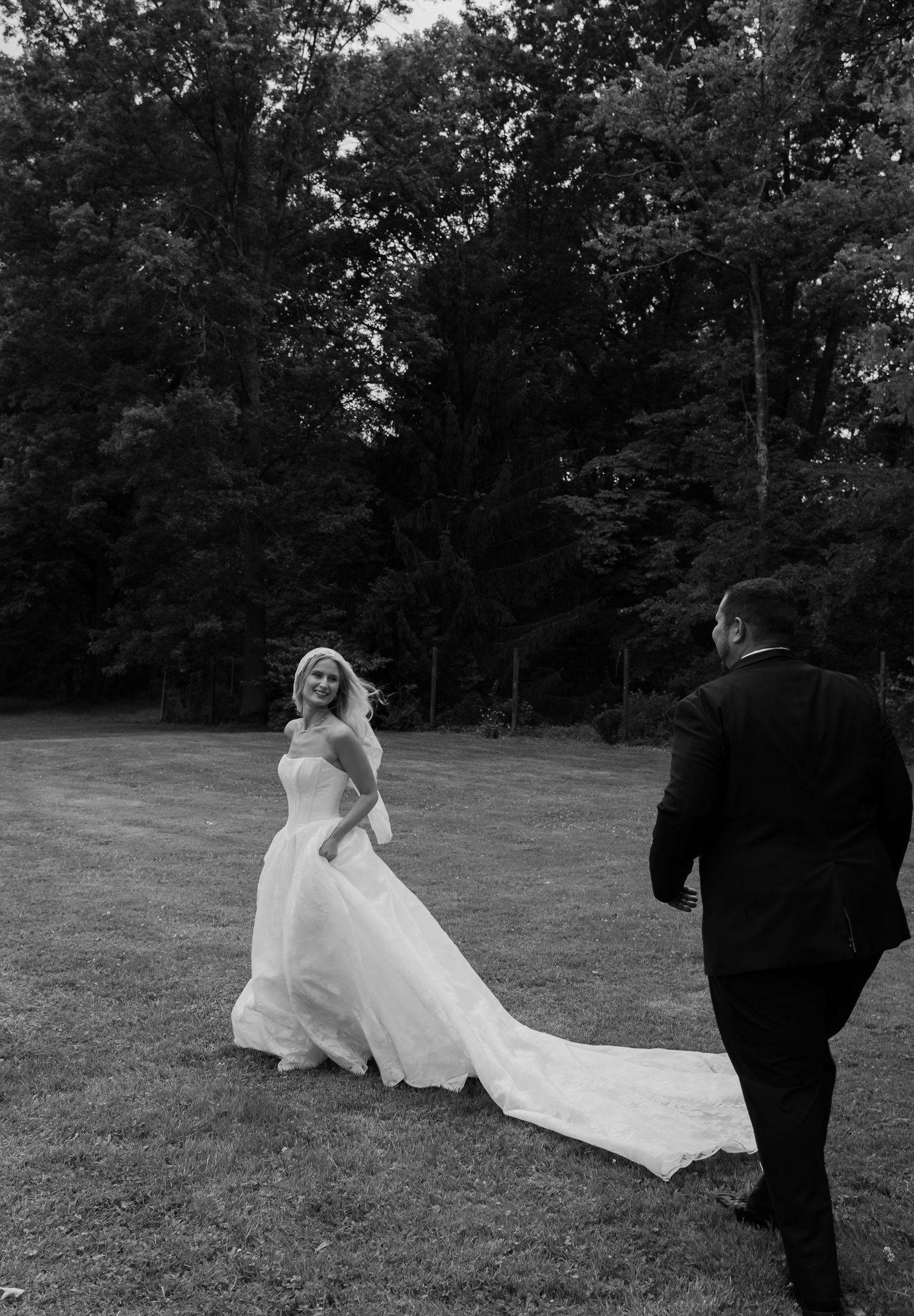 Black and white photo of bride in flowing gown with train smiling at groom outdoors.