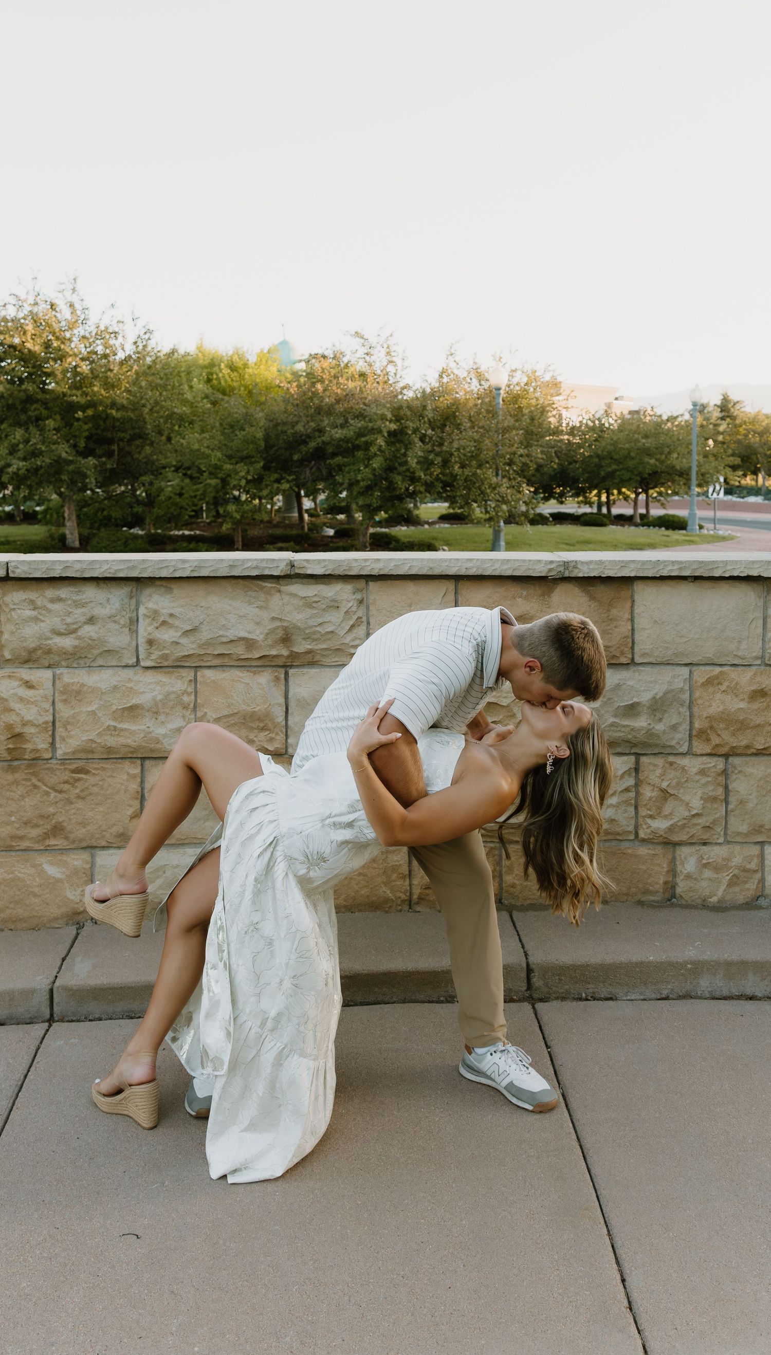 A couple shares a romantic dipping pose against a stone wall with lush greenery in the background.