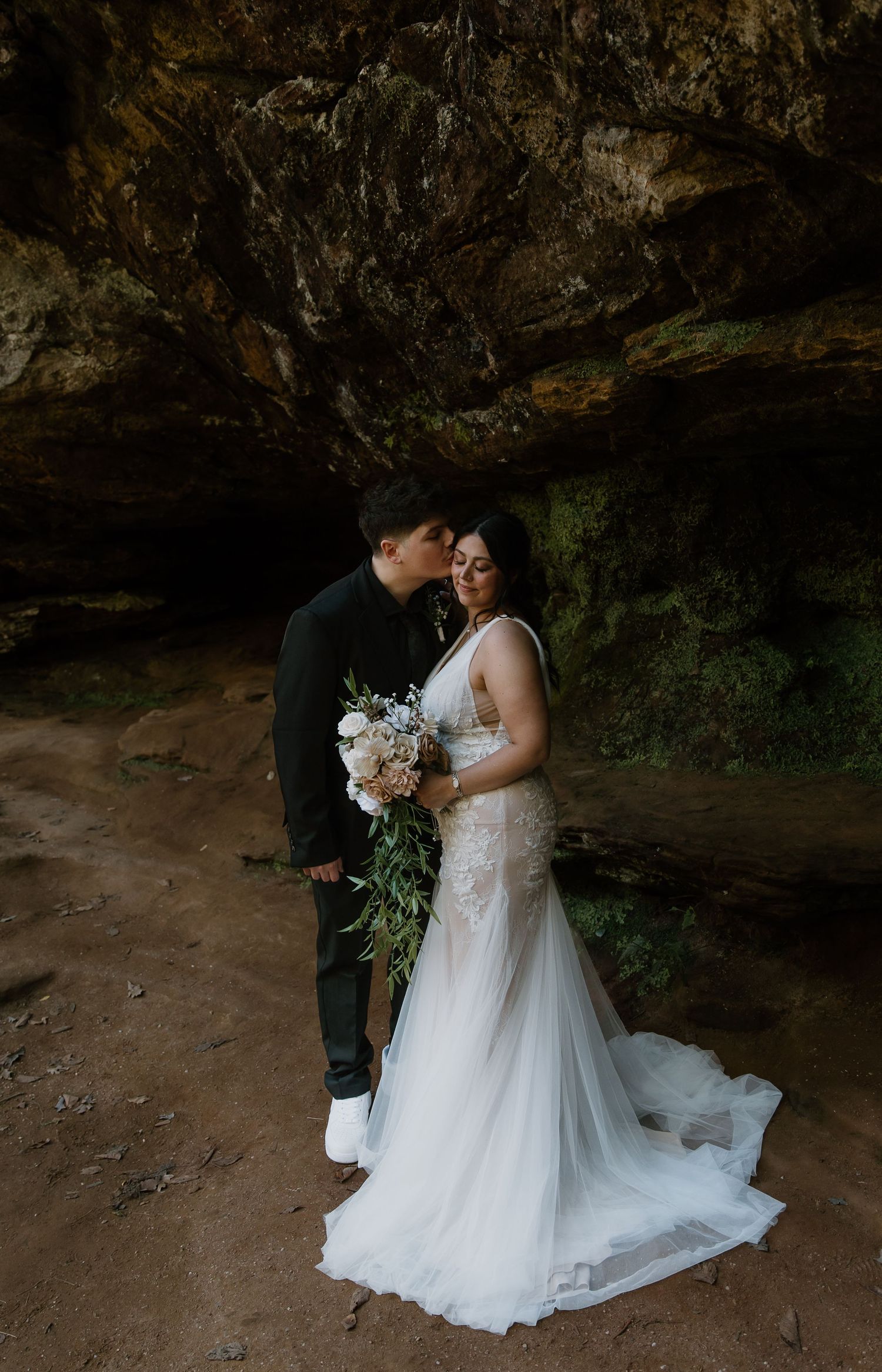 A groom kisses his bride on the forehead in a cave, her lace gown and bouquet glowing softly in the dim natural light.
