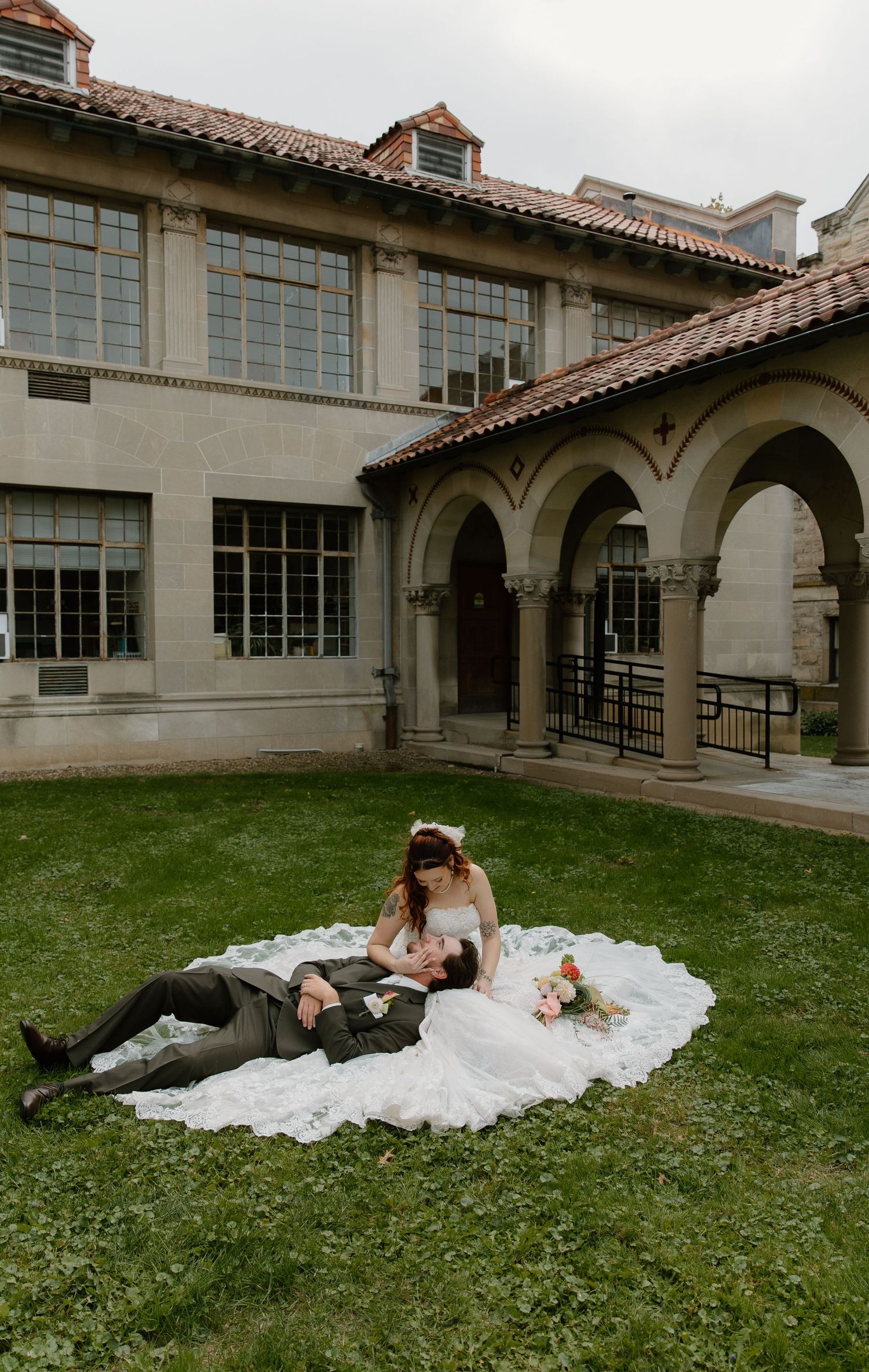 Bride and groom posing on grass outside elegant stone building with arched colonnade on their wedding day.