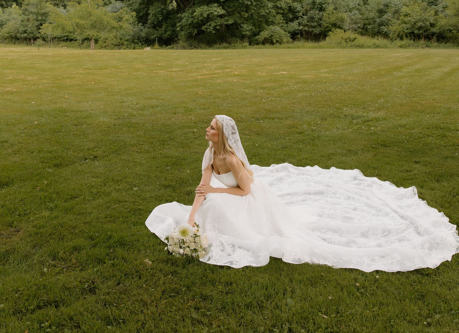 Bride in white gown and veil sits gracefully on green grass, holding a bouquet, with trees in the background.