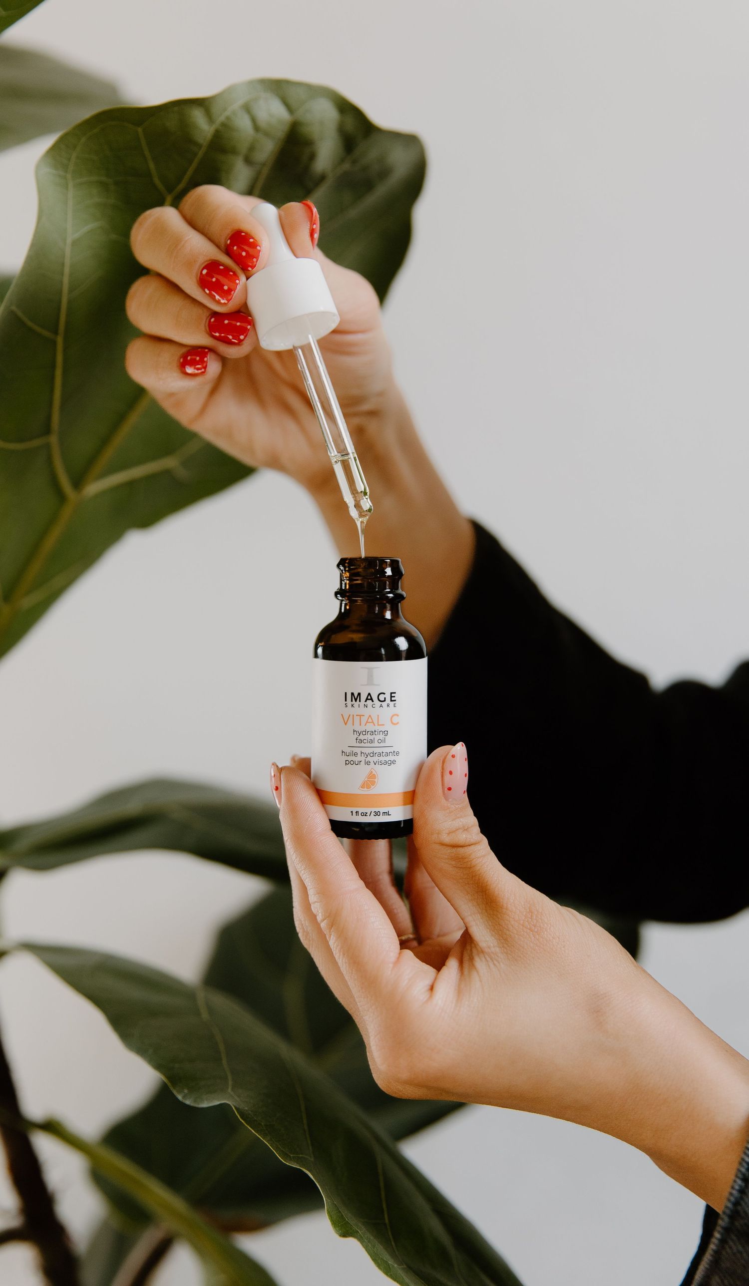 Hands holding IMAGE Skincare facial serum dropper bottle with red manicure against tropical plant background.