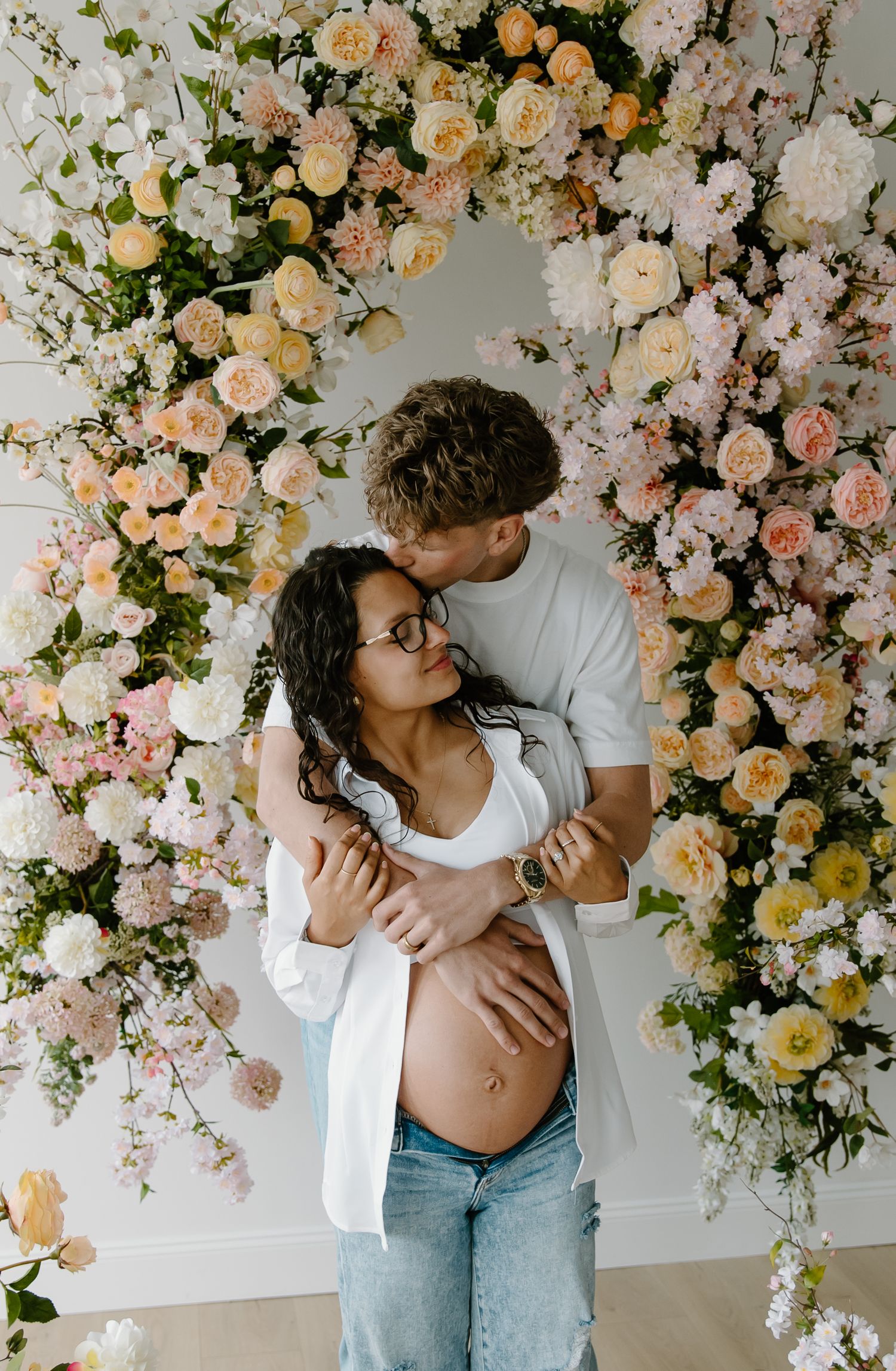Couple embracing in front of a floral arch with pink and peach roses, woman showing pregnant belly in a bright studio.