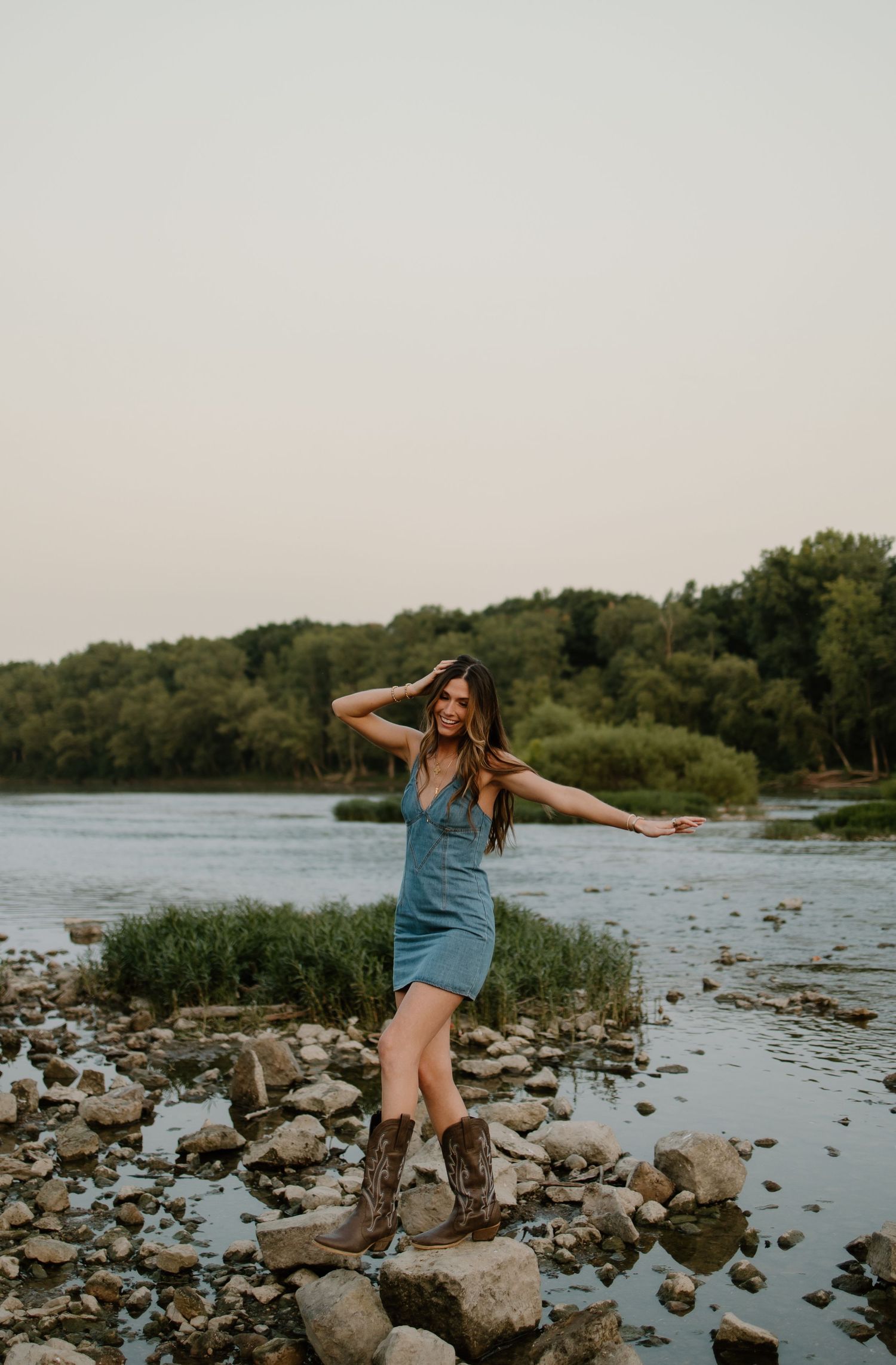 Woman in blue dress and cowboy boots posing on rocky riverbank with lush green trees in background at dusk.