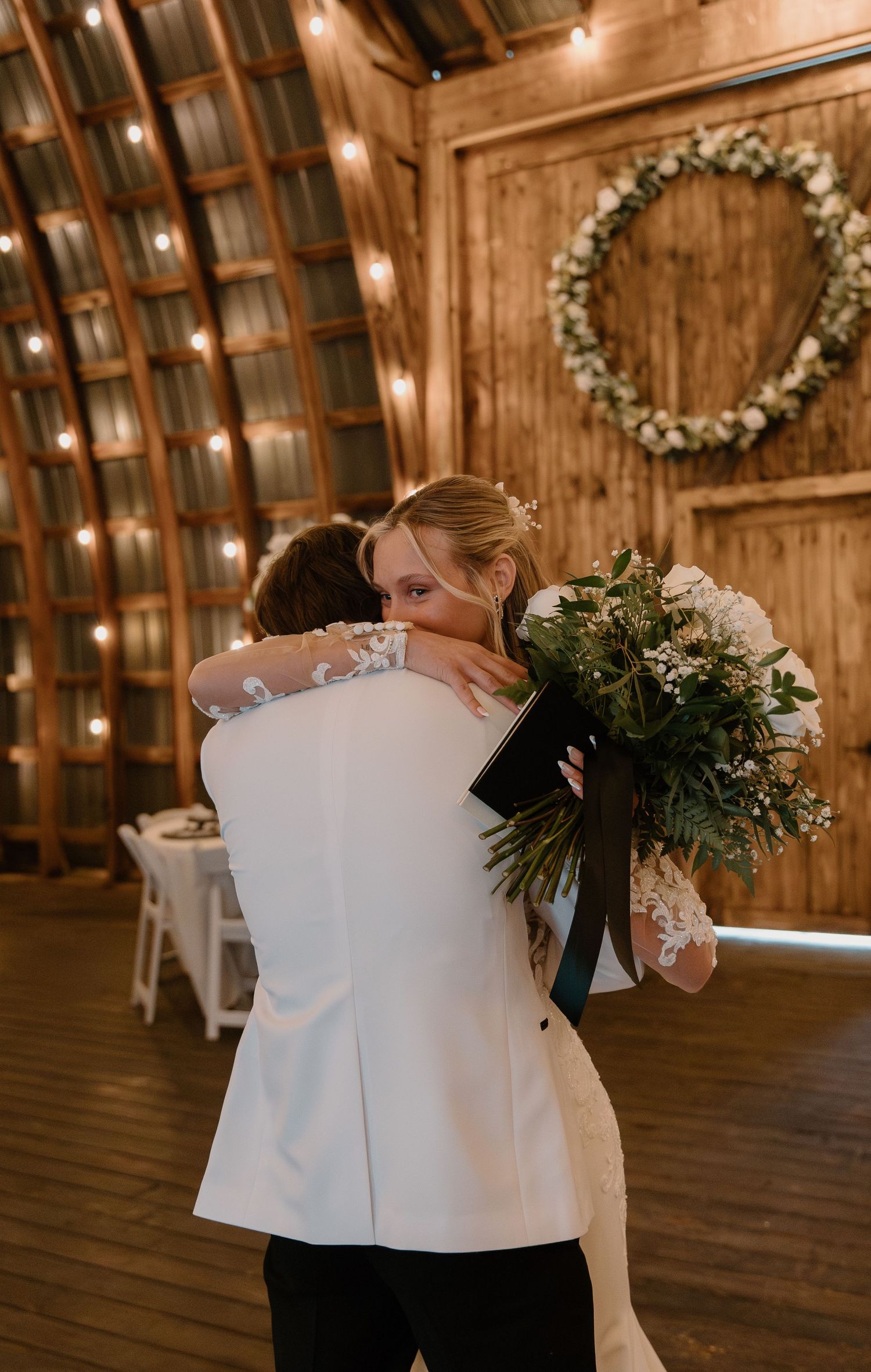 Bride and groom share an emotional first look hug inside a rustic barn wedding venue with string lights.