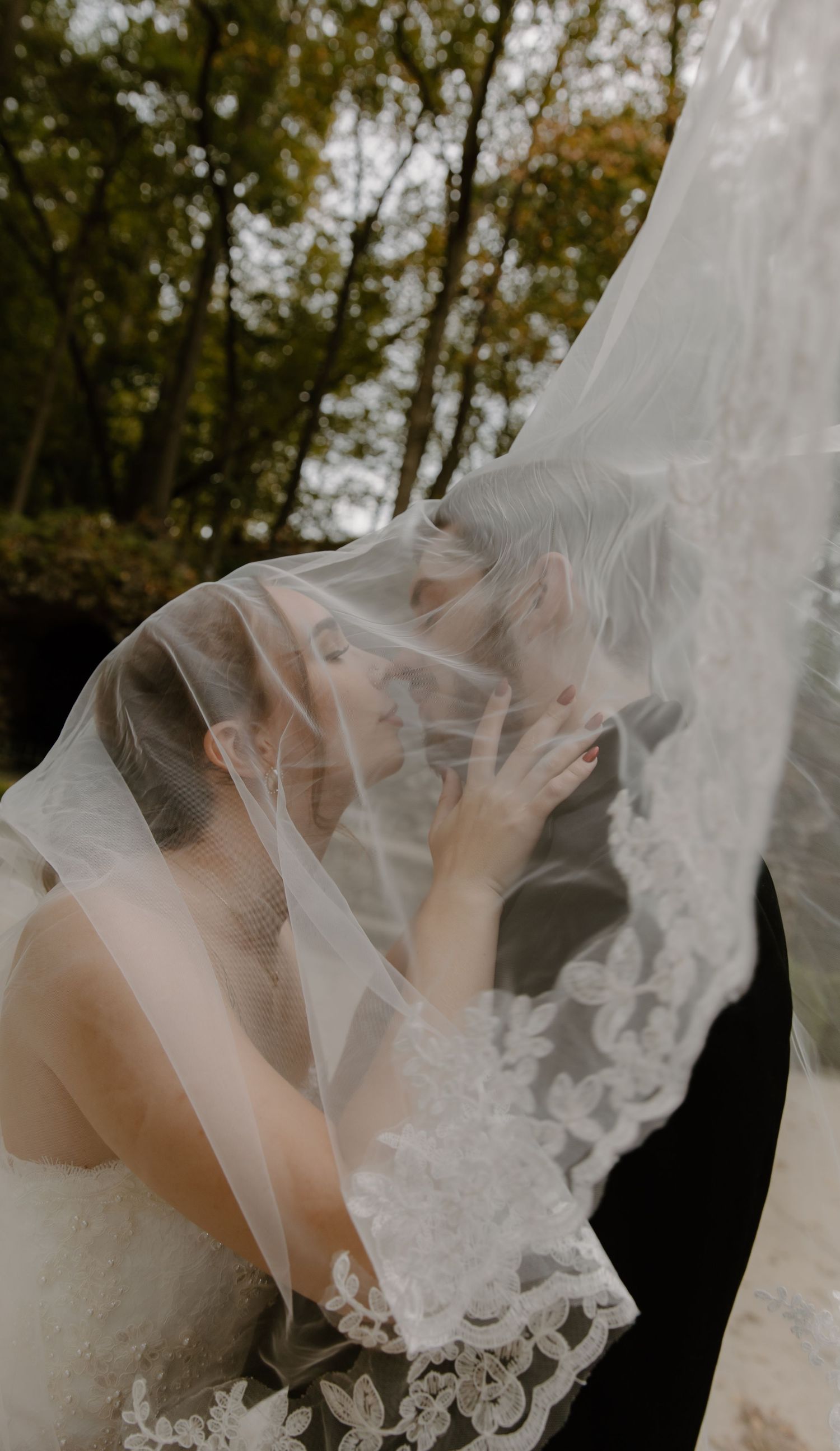 A delicate lace wedding veil flows gracefully in front of autumn trees creating an ethereal bridal portrait.