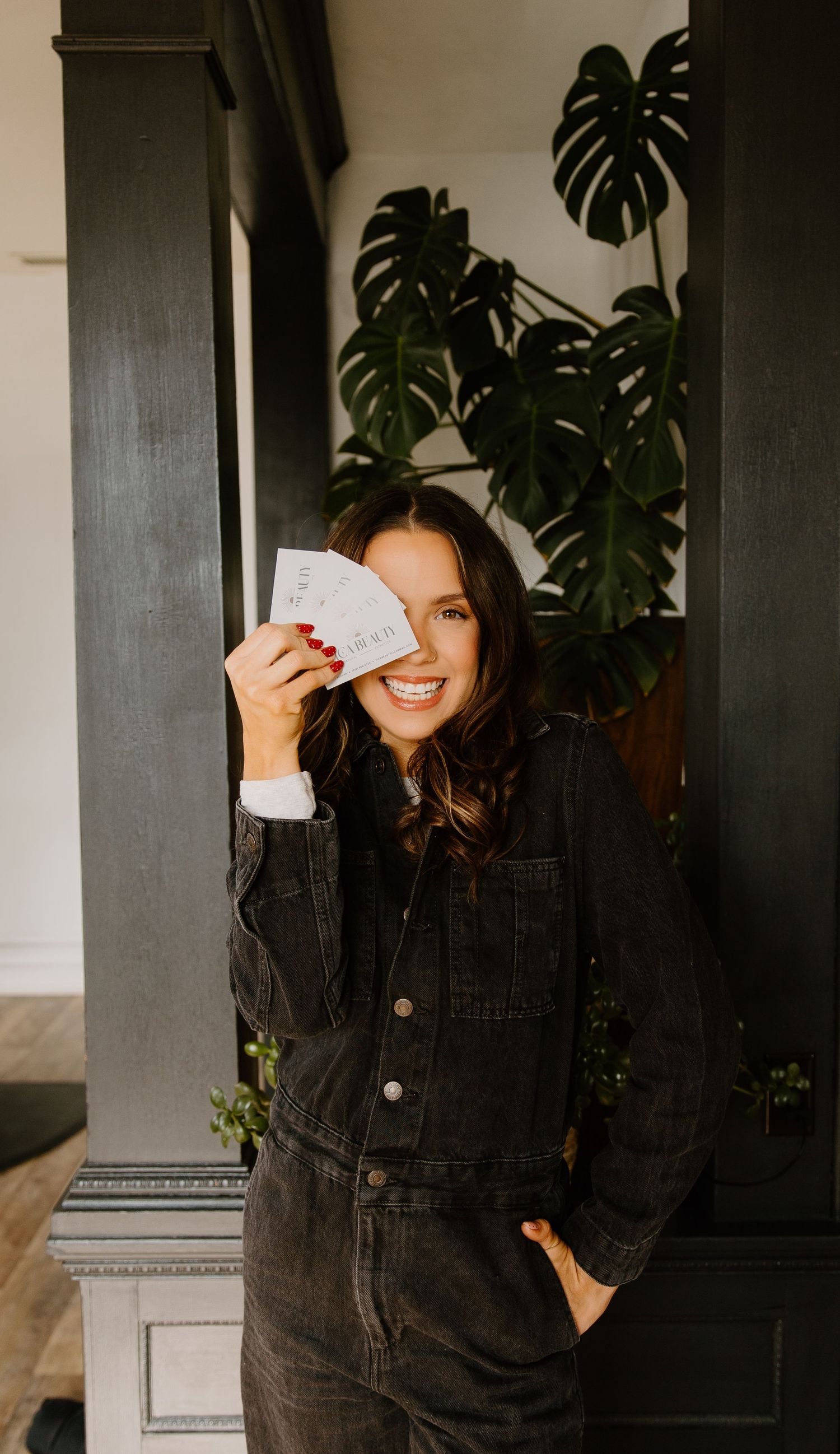 Smiling woman in black denim jumpsuit holding playing cards near her face, standing by dark pillars with monstera plant.