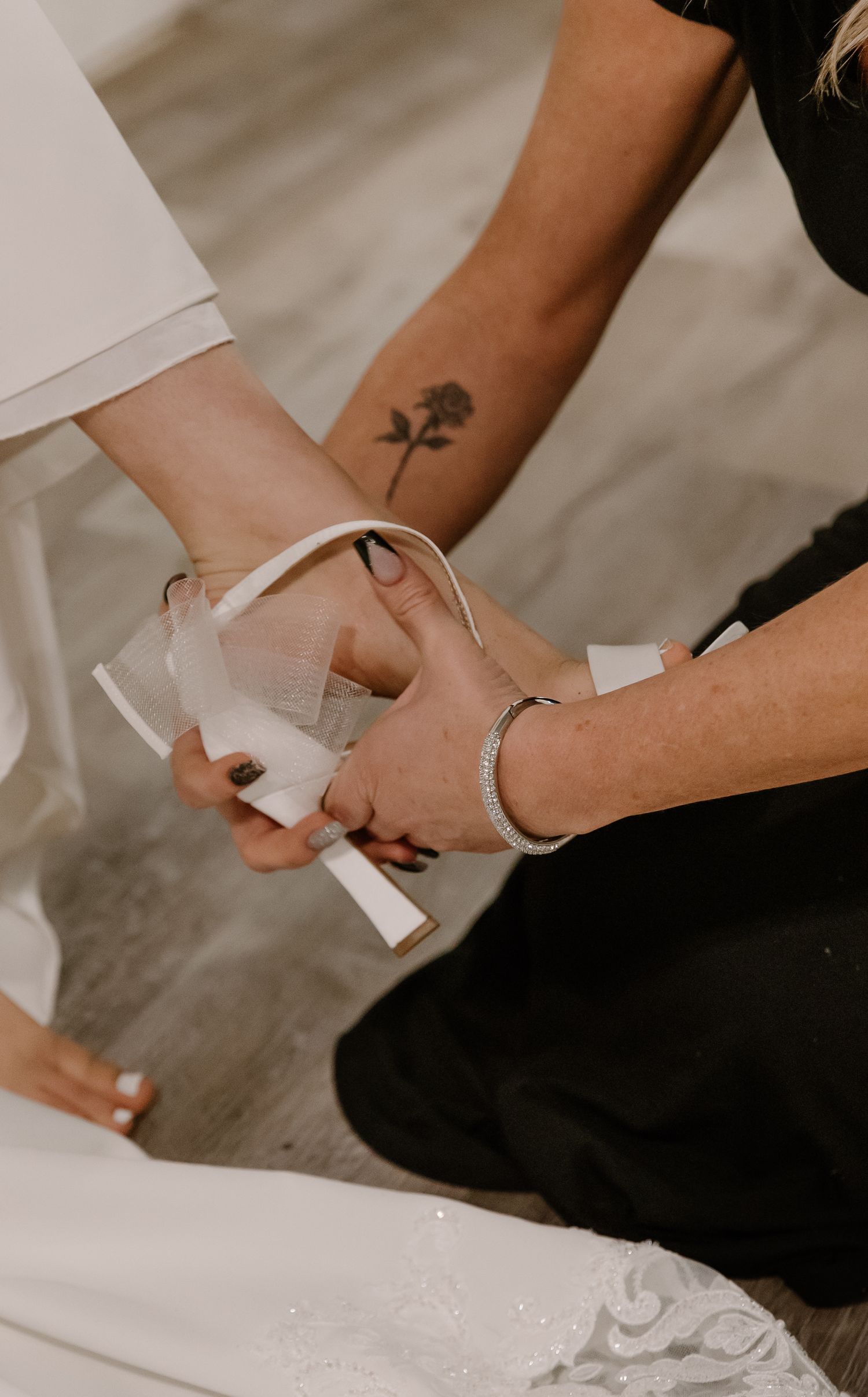 Person with rose tattoo helping bride put on white heeled sandal during wedding preparation.