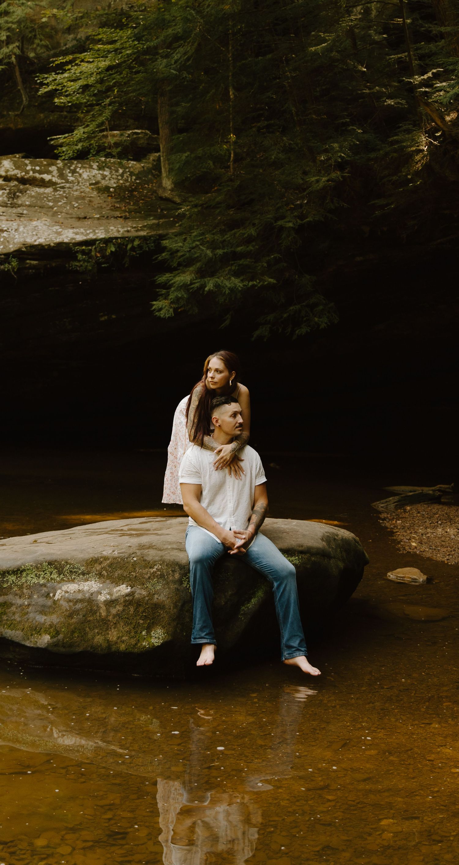 A person sits peacefully on a large rock by a serene stream in a wooded forest setting.