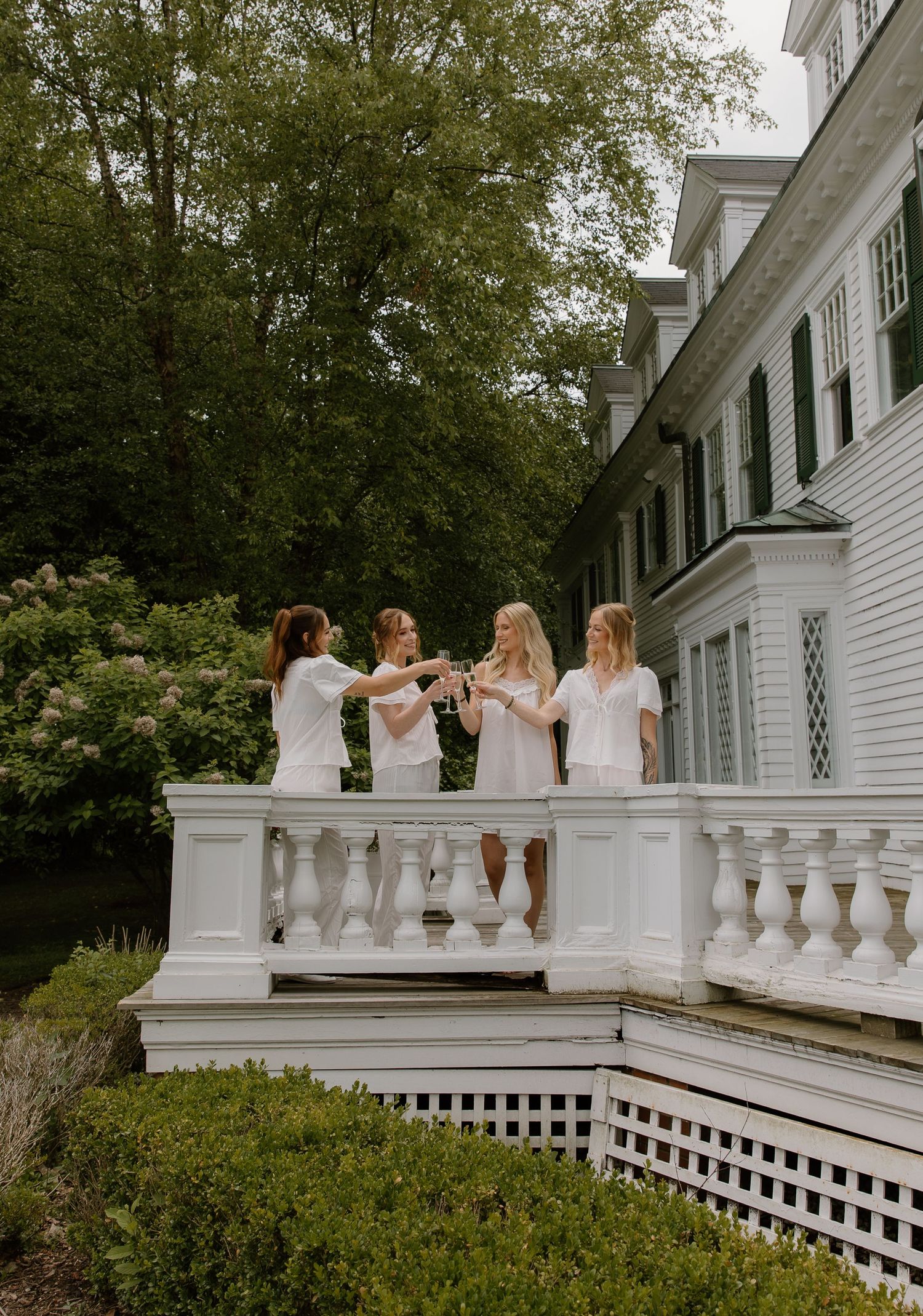 Four bridesmaids in white dresses toasting champagne on a white balcony of an elegant estate.