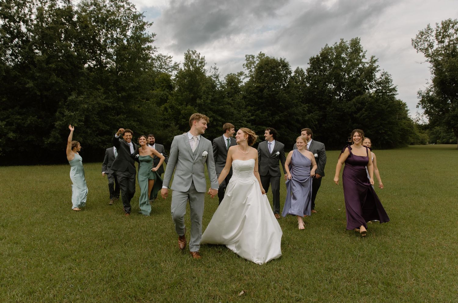 Wedding party walking across a green lawn with lush trees, bride in white gown holding groom's hand, bridesmaids in purple dresses.
