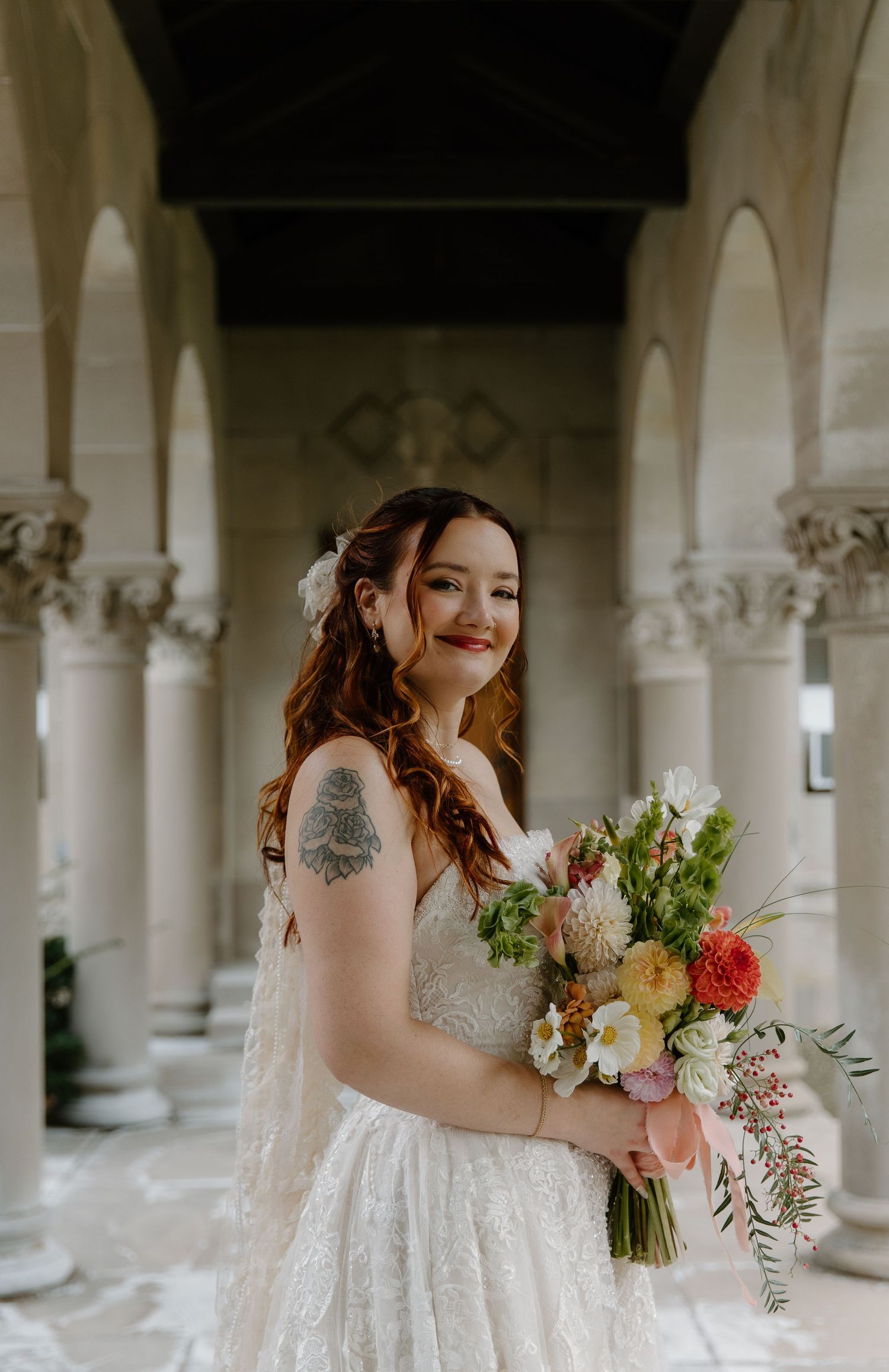 Bride in lace wedding dress holding colorful bouquet, smiling among elegant stone columns.