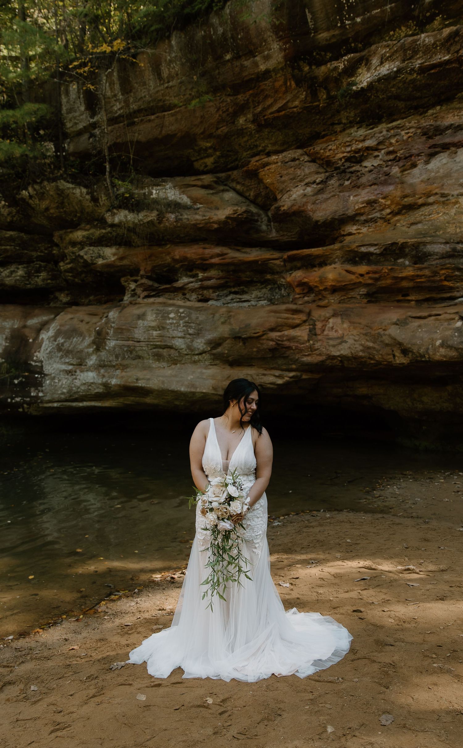 Bride in white gown holds cascading floral bouquet before sandstone cliffs and serene pool in nature.