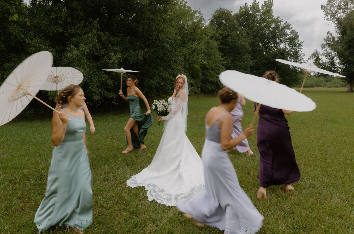 Bride and bridesmaids in colorful gowns walking outdoors holding white parasols on a cloudy wedding day.