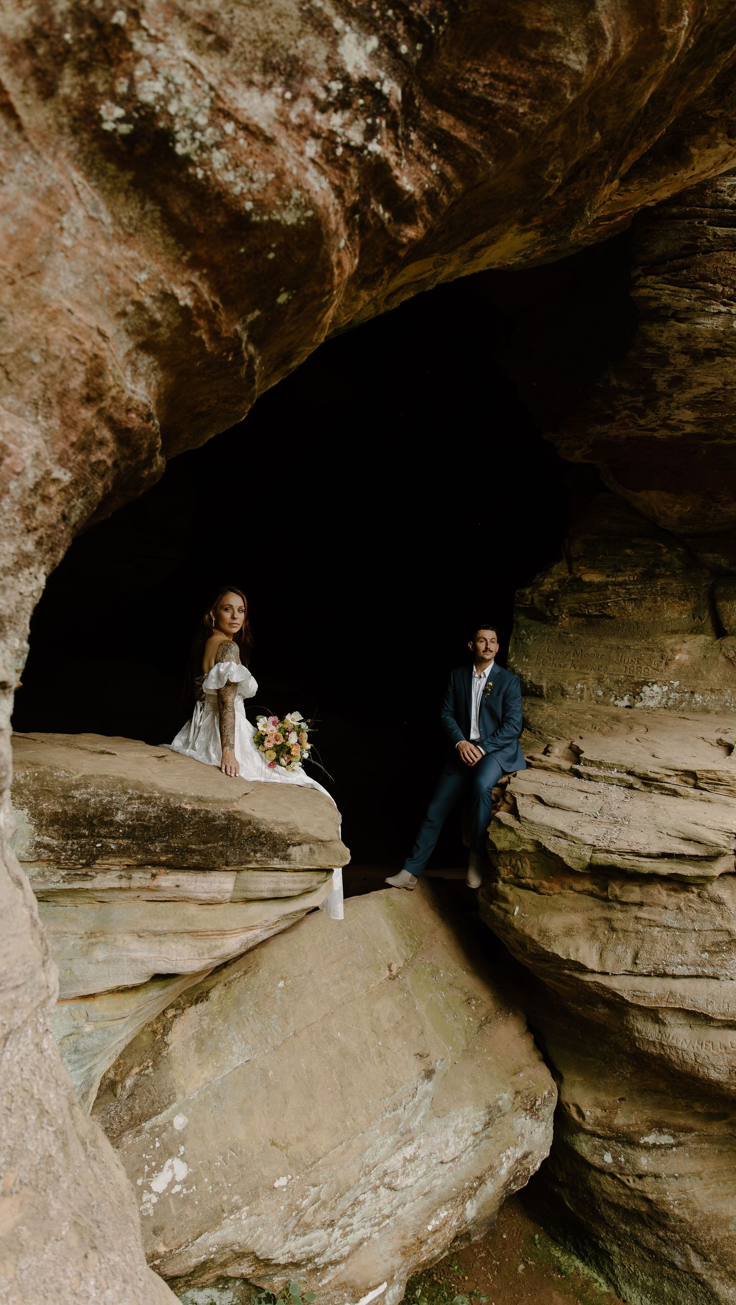 A couple in wedding attire poses together in a dramatic rocky cave formation for their wedding photos.