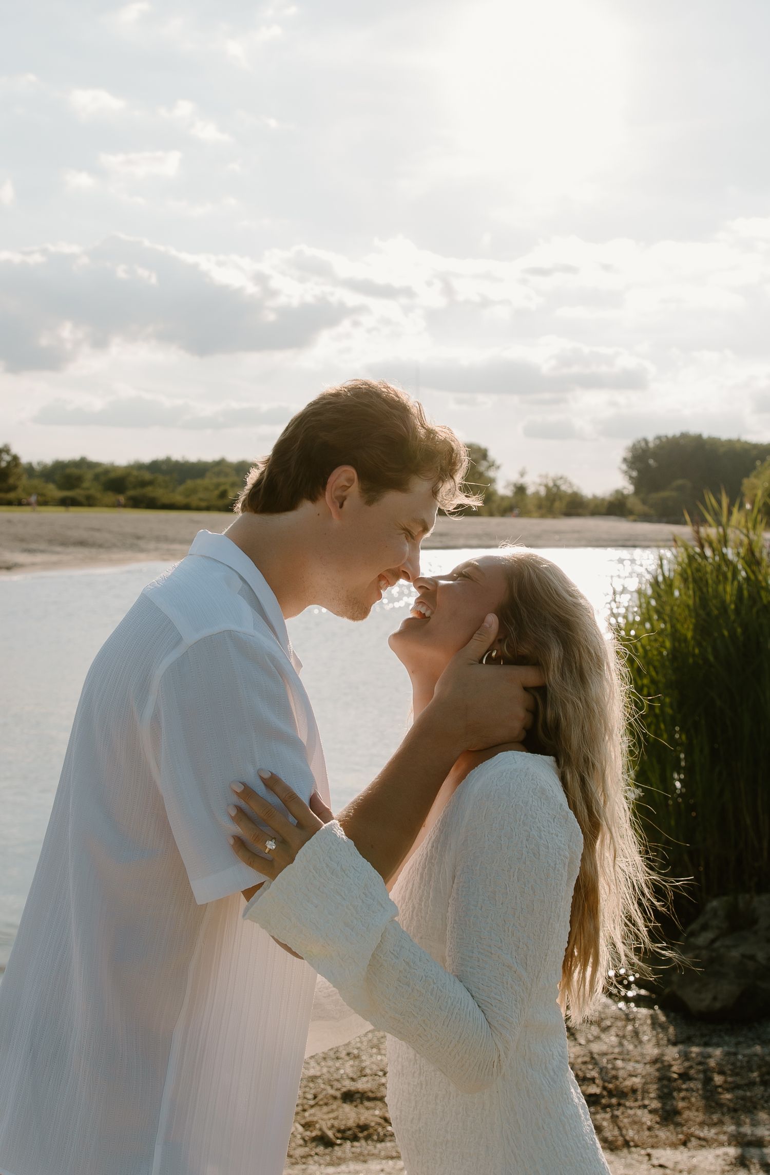A couple in white outfits sharing a romantic moment by a sunlit lake shore surrounded by lush greenery.