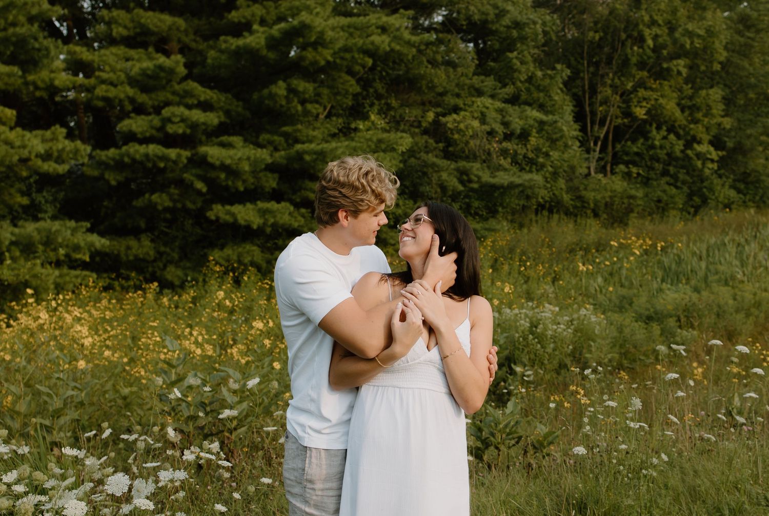 Couple embracing in a wildflower meadow, woman in white dress, surrounded by lush green trees on a warm summer day.