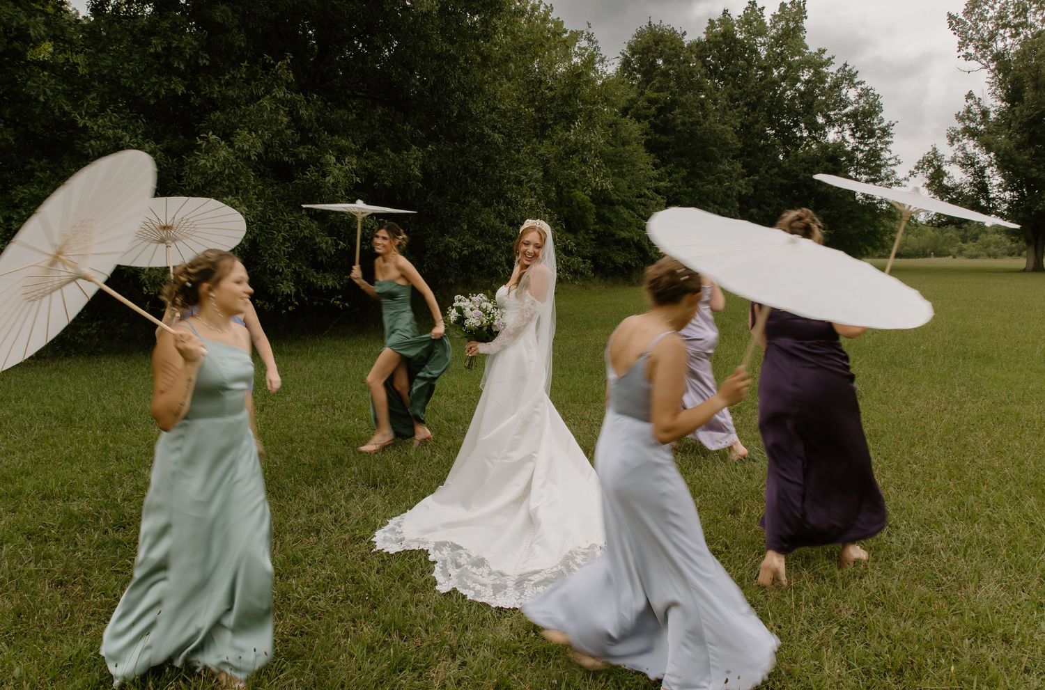 Bride and bridesmaids in colorful gowns twirling white parasols on green grass at an outdoor wedding.
