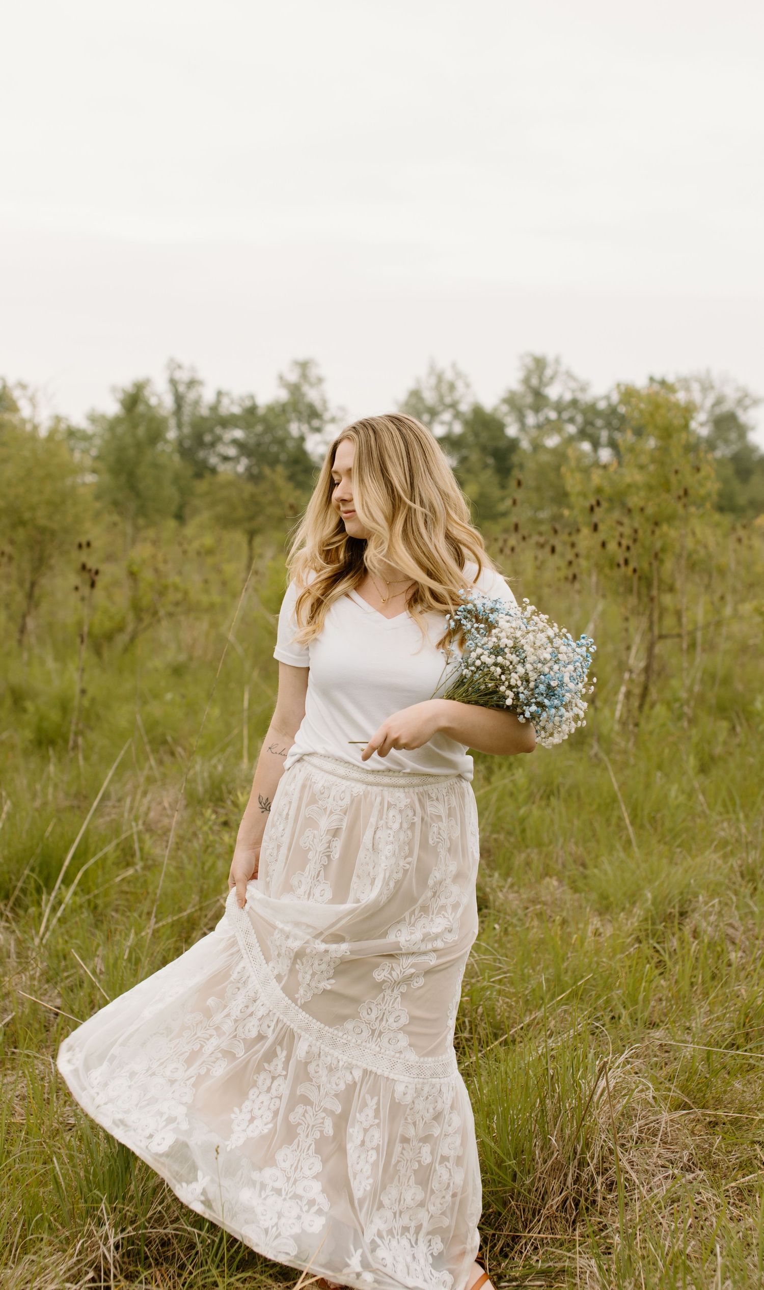 Woman in flowing white lace skirt and top holds wildflowers while standing in a sunlit meadow in ohio.