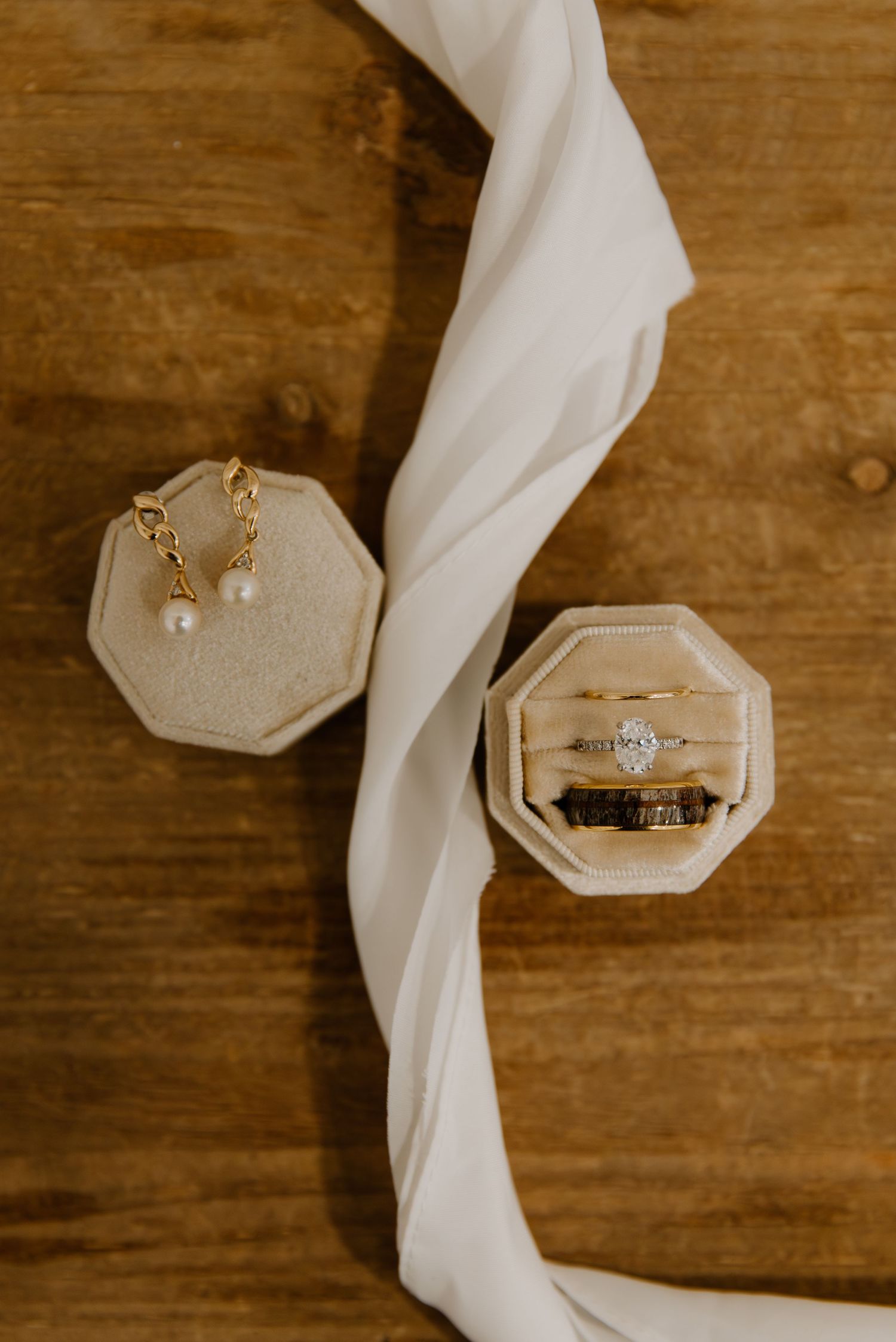Wedding rings and pearl earrings displayed in octagonal velvet ring boxes with white ribbon on wooden surface.
