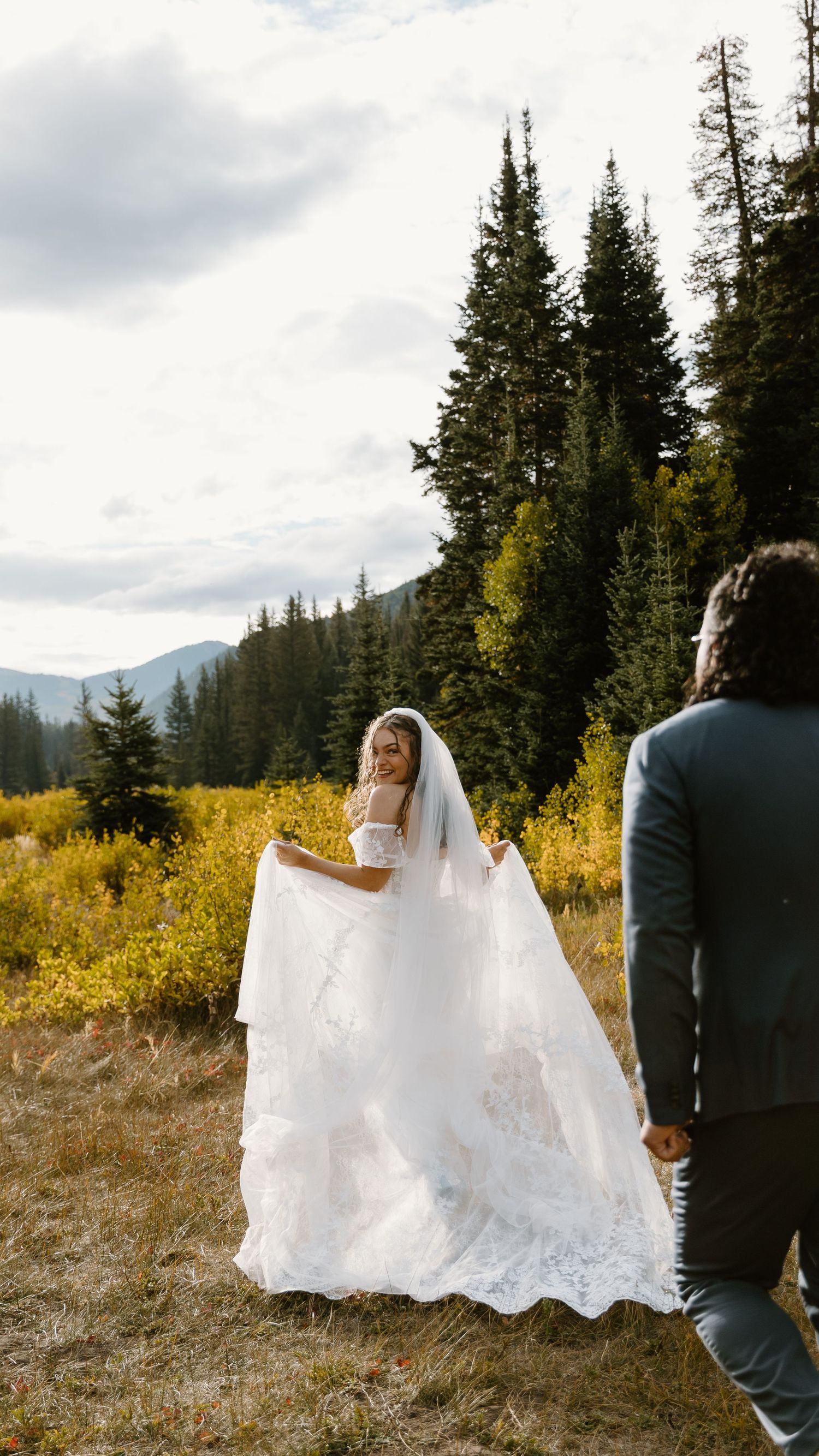 A wedding couple walks through a scenic mountain meadow with evergreen trees and golden foliage.