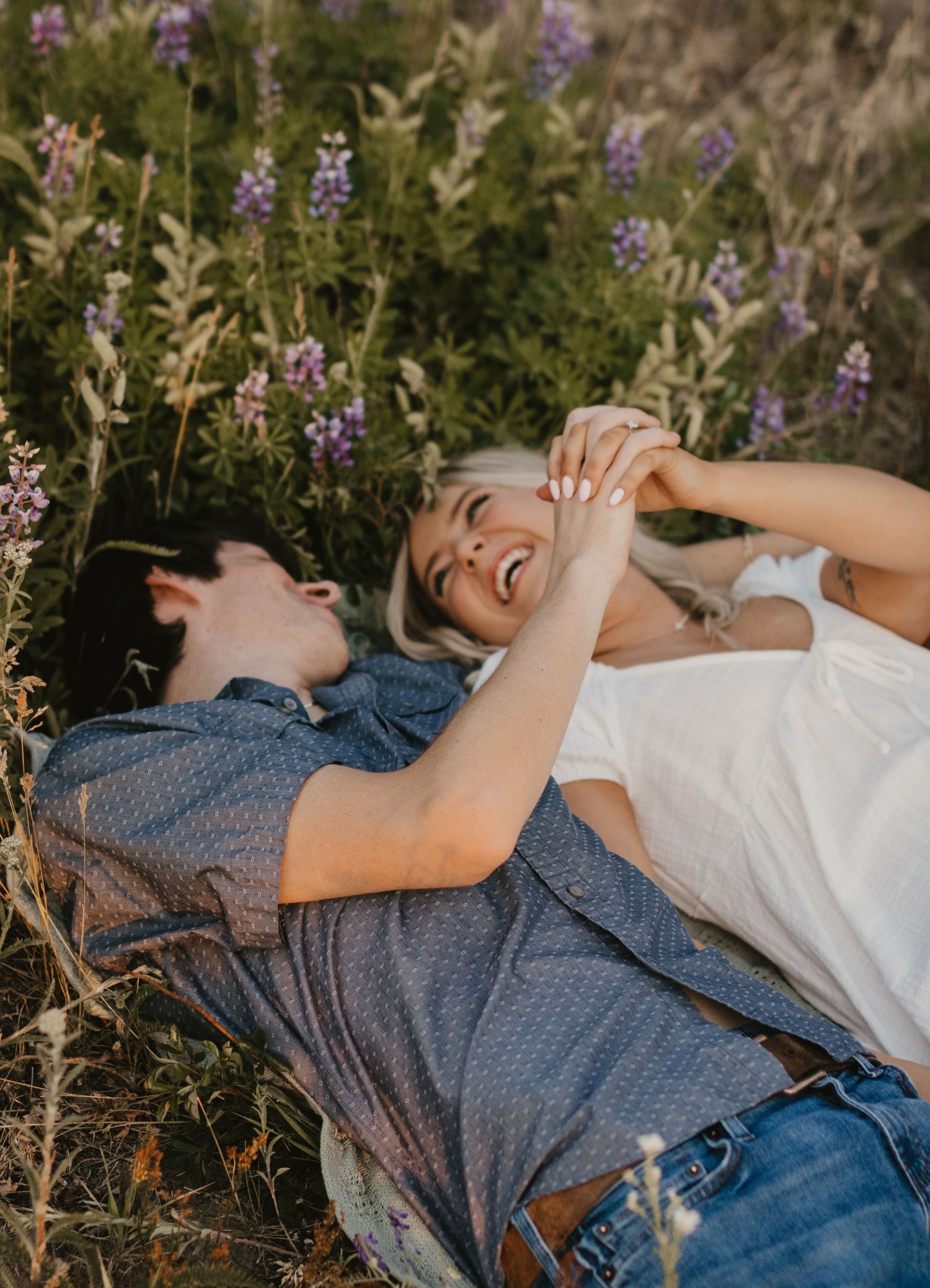 A couple lies in a field of purple wildflowers, sharing laughter and joy.