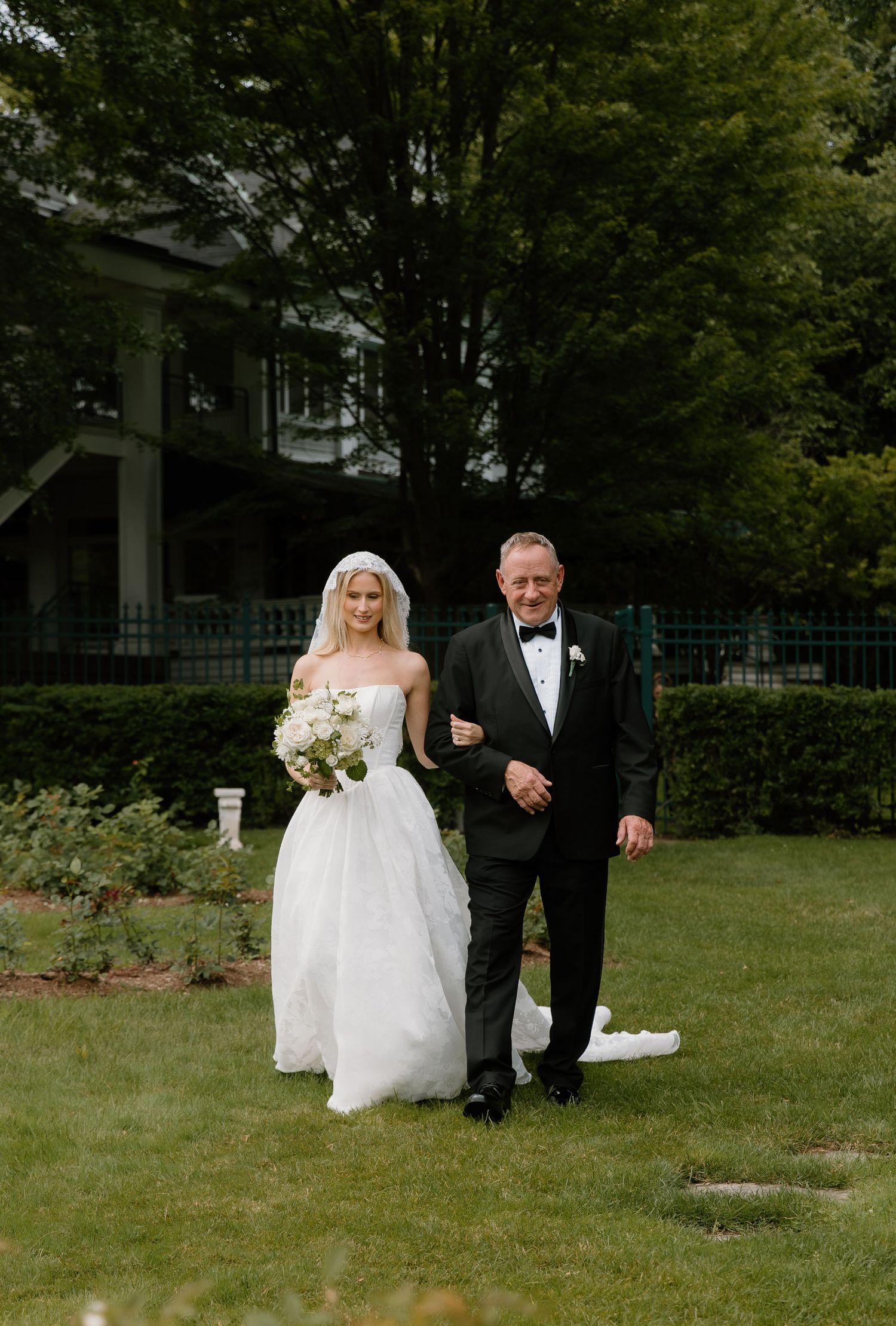 Bride in white strapless gown with veil walks arm-in-arm with father in tuxedo on green lawn.