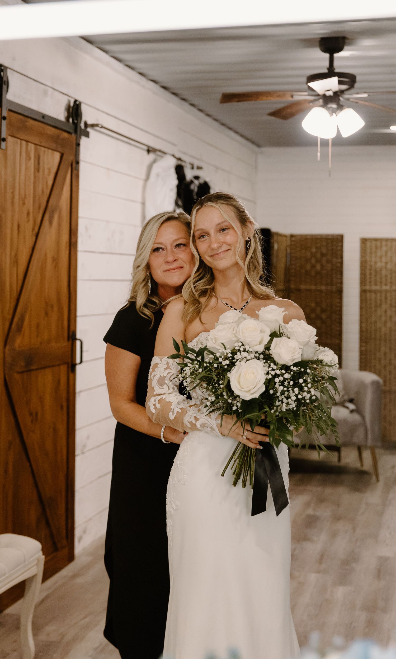 Bride in white lace gown holds white rose bouquet with woman in black dress in rustic barn venue.
