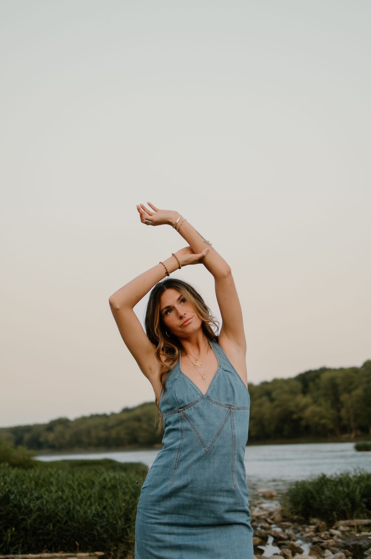 Woman in denim dress poses with arms raised at golden hour near a serene river with lush greenery.