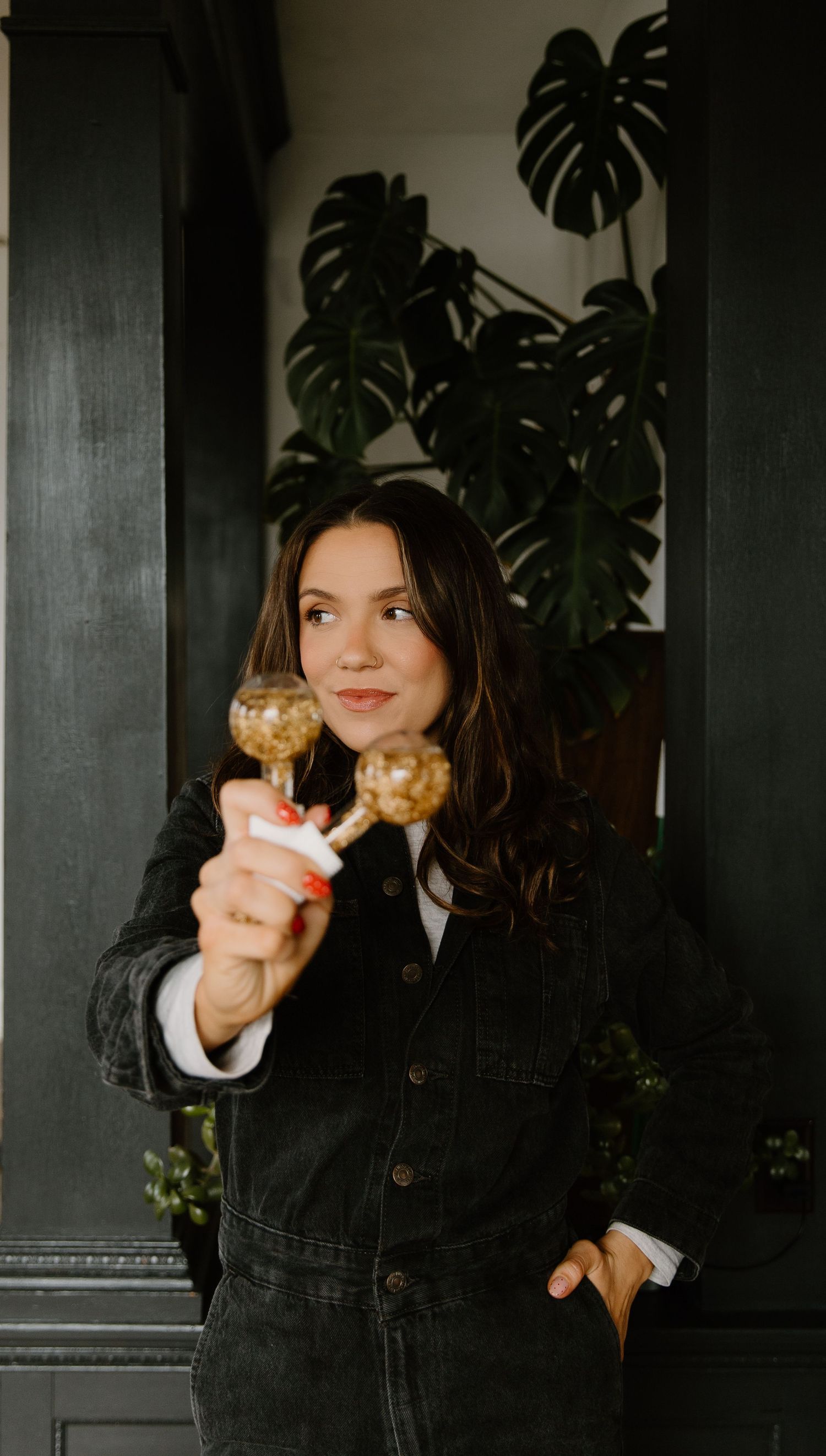 Woman in black denim outfit holding cake pops, posing indoors with monstera plant backdrop.