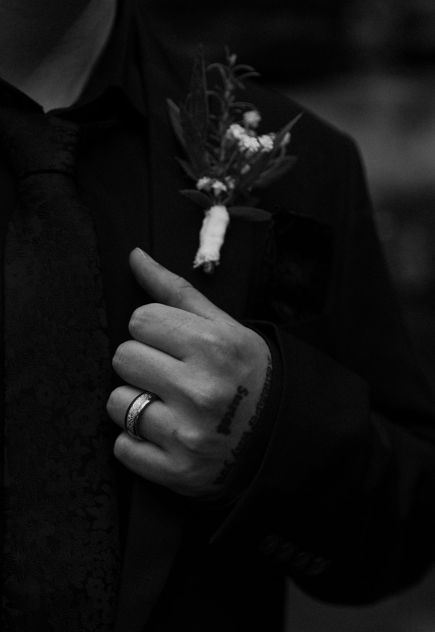 Black and white photo of a groom in a suit adjusting his floral boutonniere, wearing a silver ring.