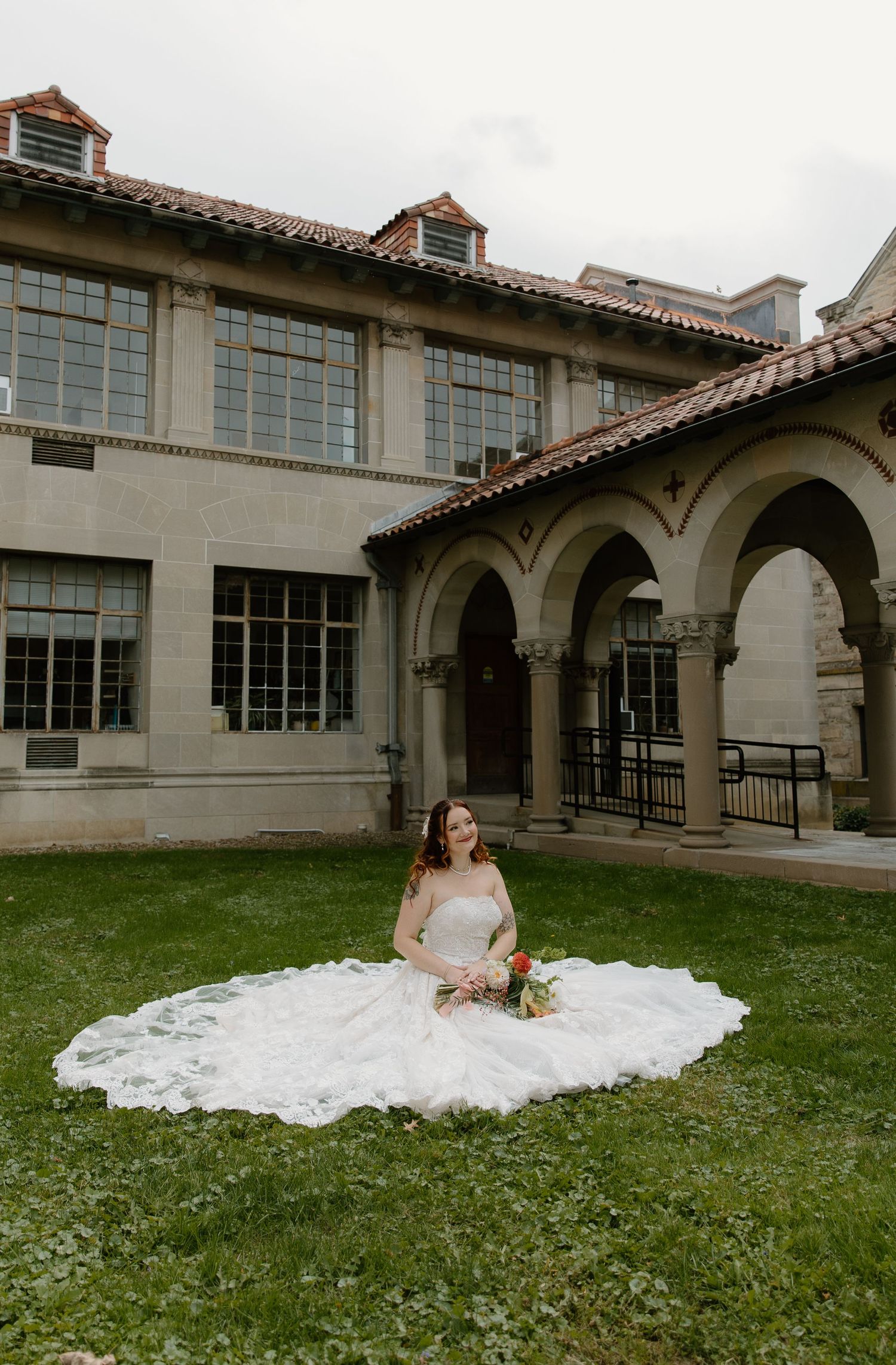 Bride in a white ball gown sits on green grass in front of an elegant stone building with arched colonnades.