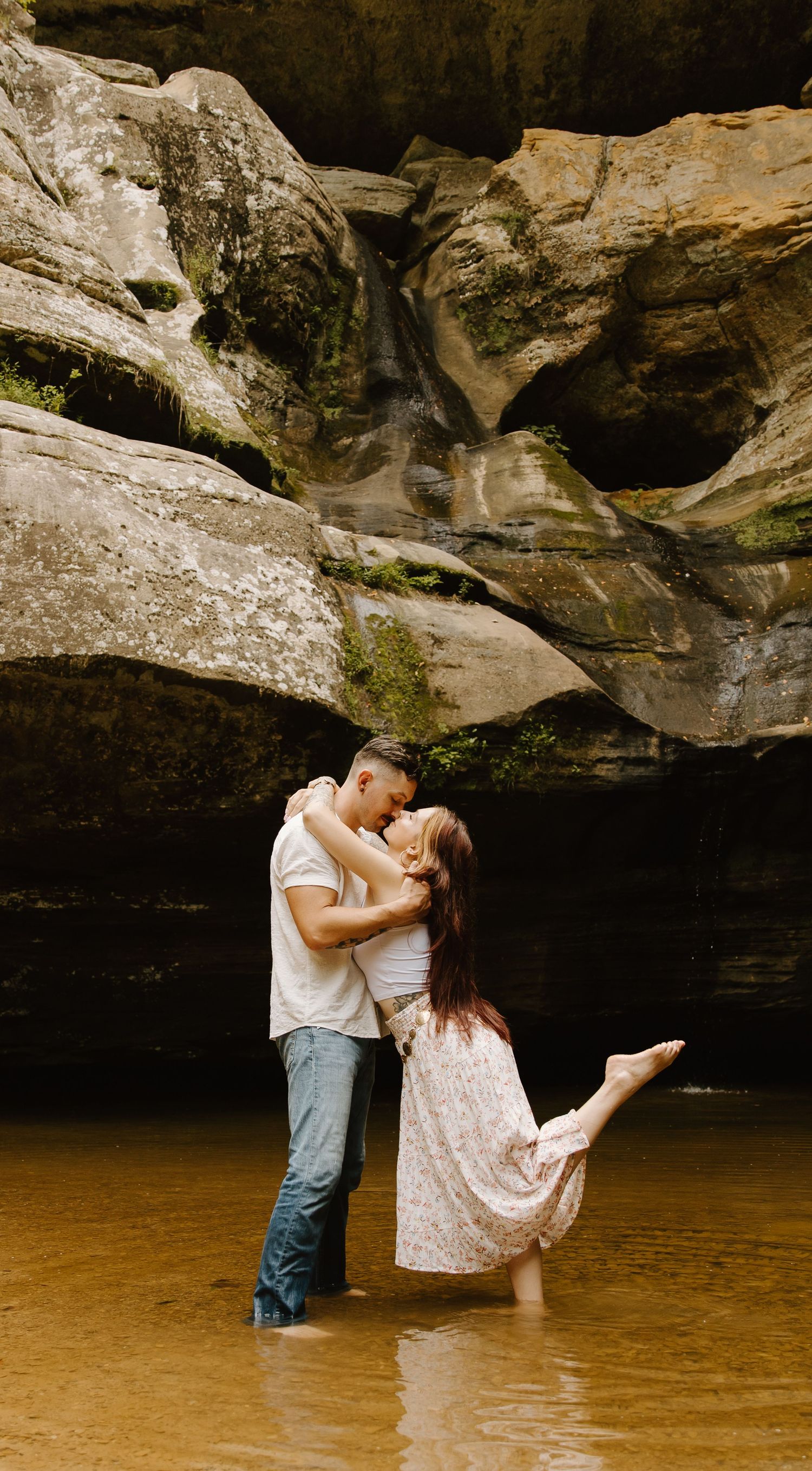 A romantic moment shared between a couple standing in shallow brown water beneath dramatic rock formations during an engagement shoot.