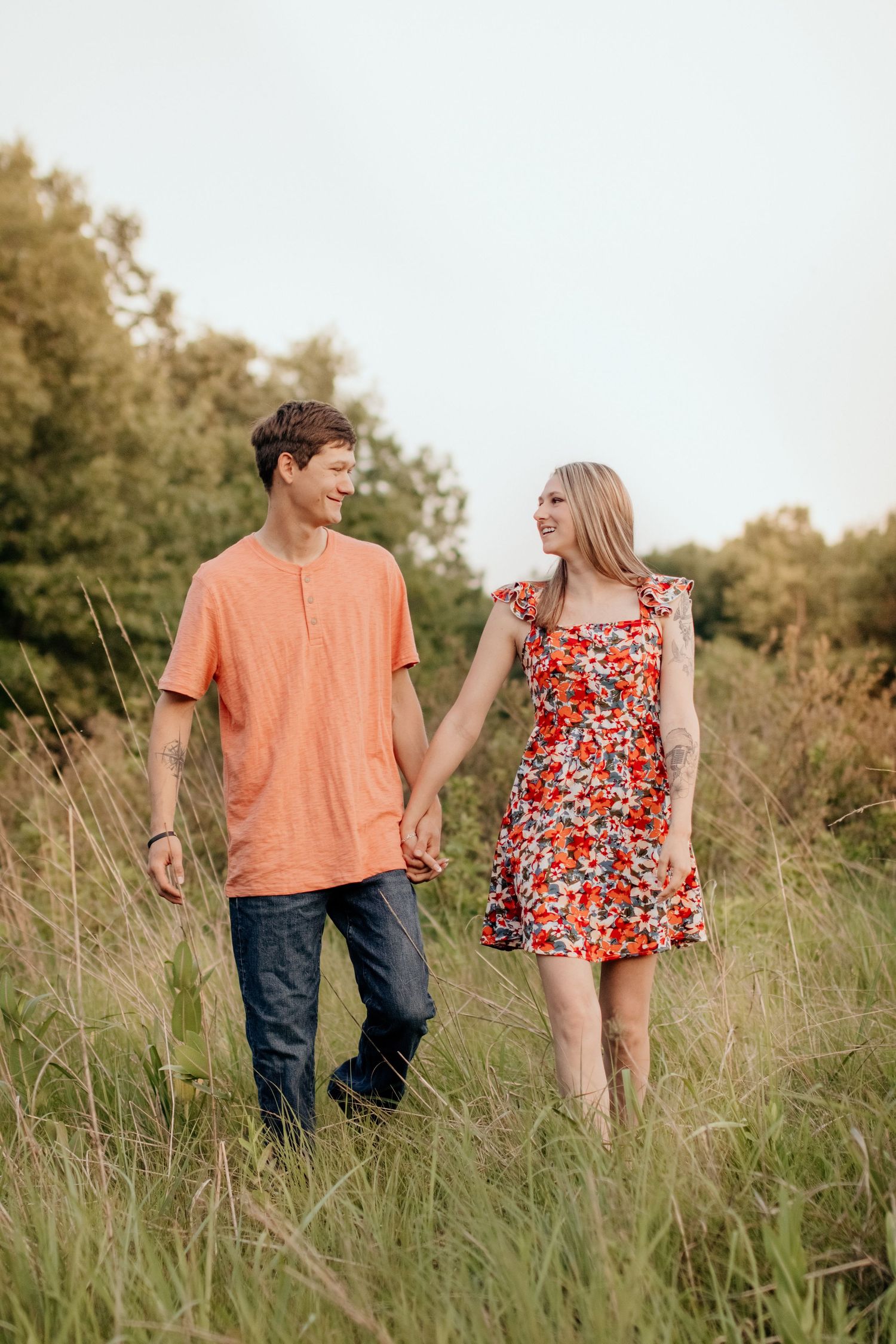 A couple walks hand in hand through tall grass wearing coordinated orange and floral outfits.