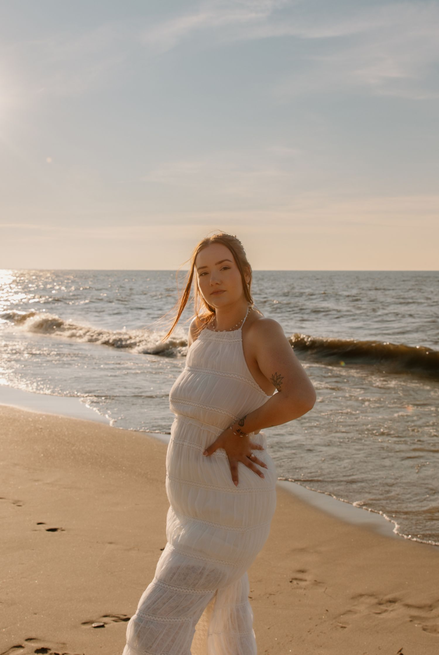 Pregnant woman in white lace dress posing on sandy beach at golden hour with ocean waves behind her.
