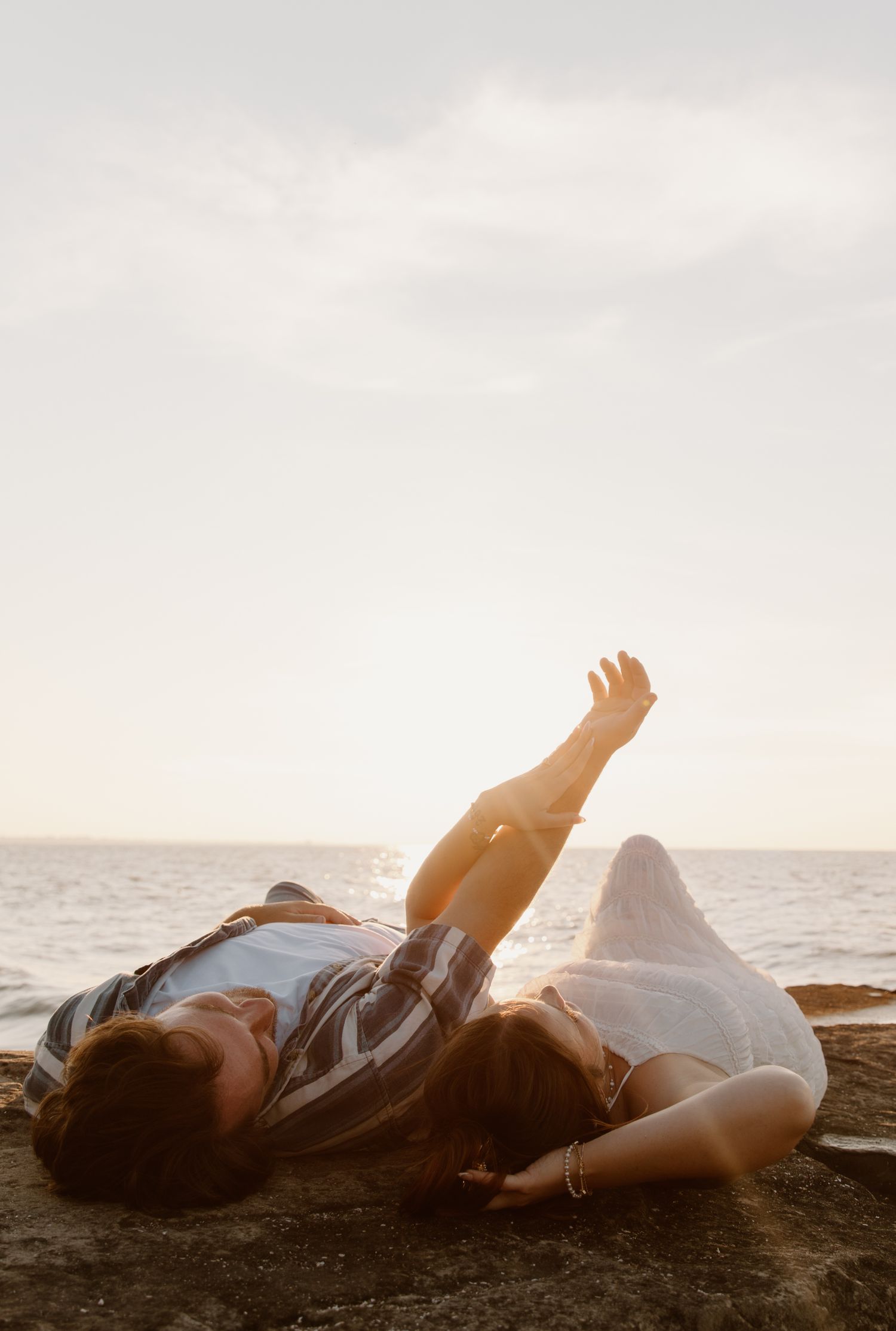 Couple lying on rocky beach at sunset, woman raising her arm toward the sky near the ocean.