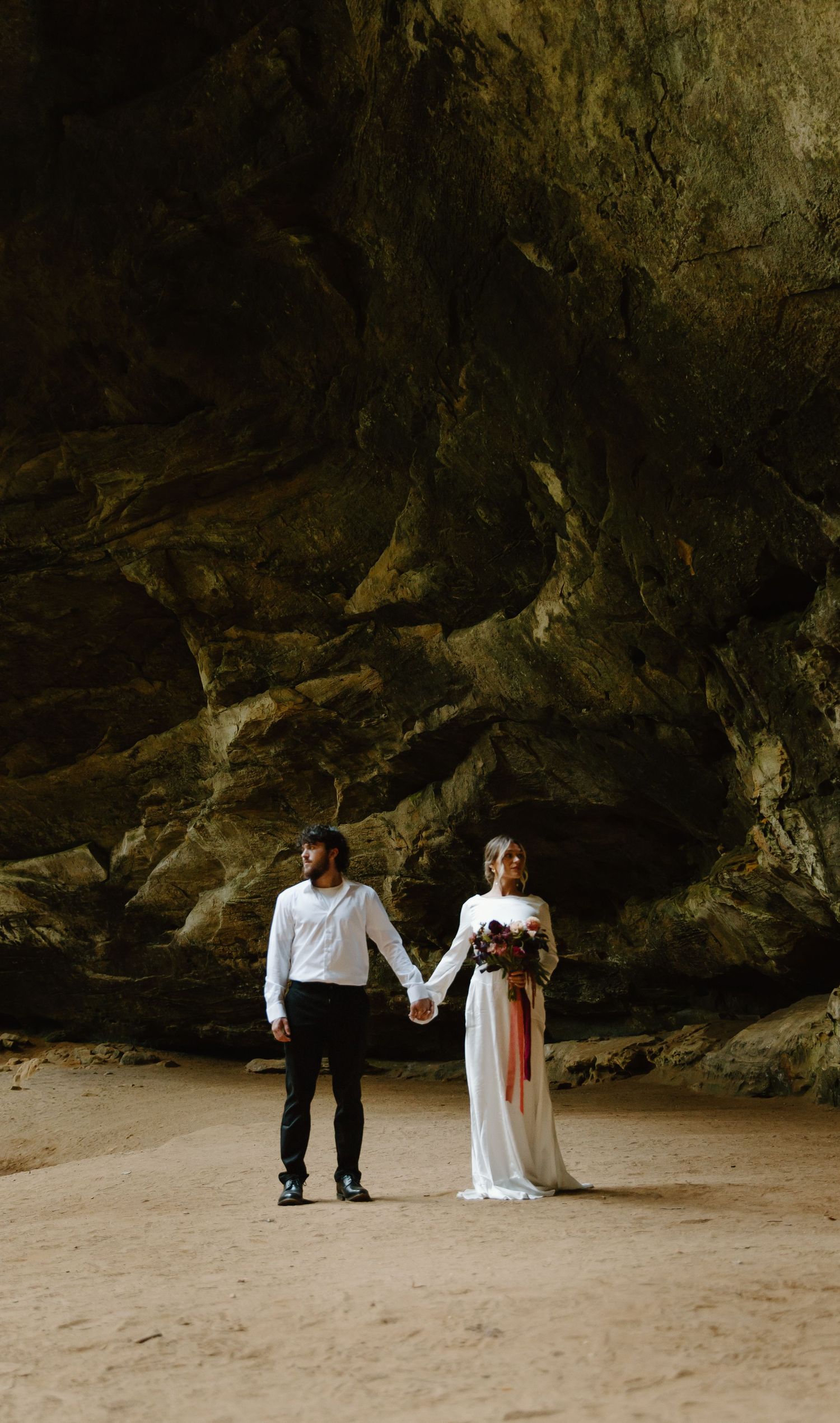 A couple in wedding attire poses under a dramatic rocky cave overhang on a sandy beach during a photoshoot.